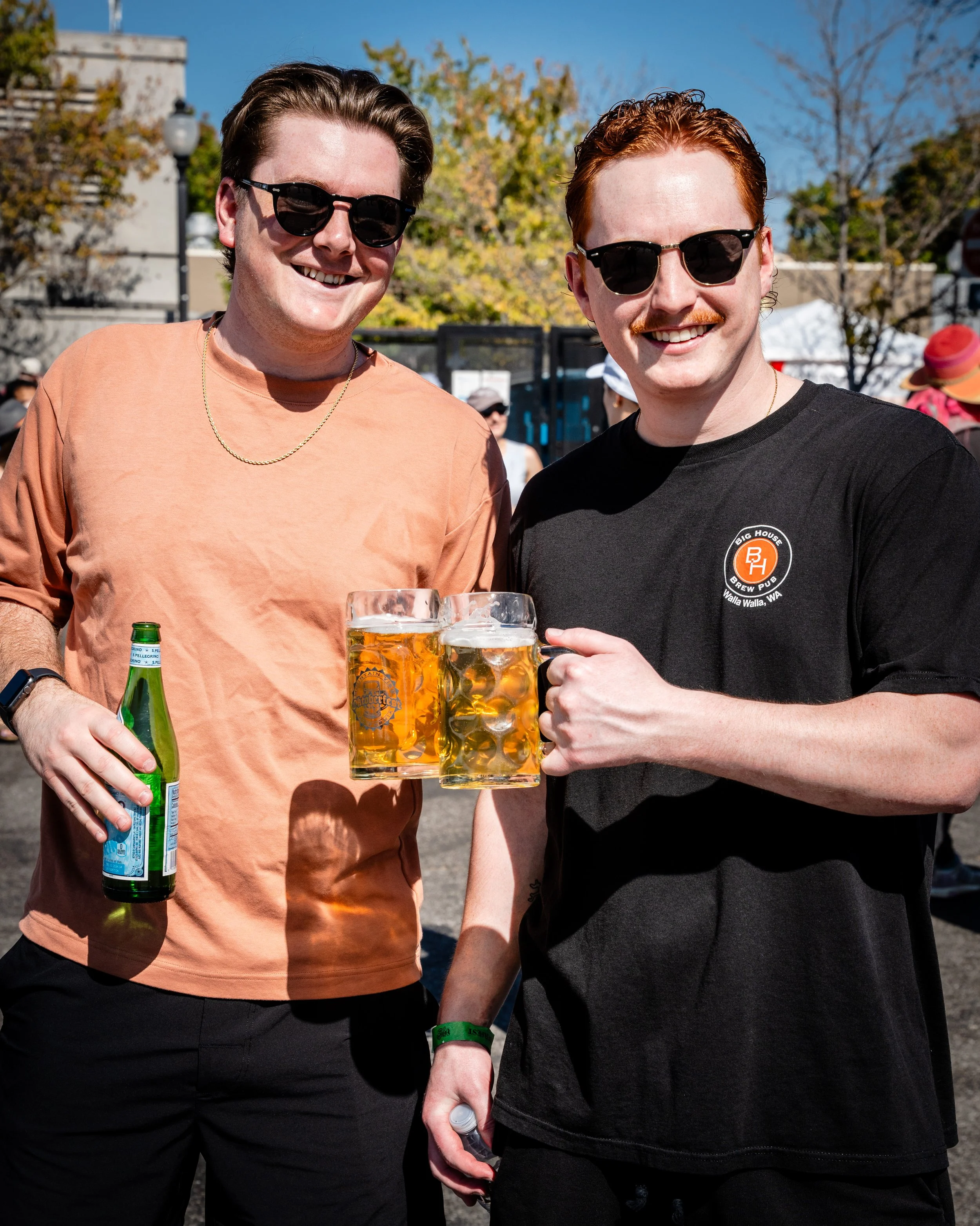 Two men smiling outdoors, wearing sunglasses, holding glasses of beer and a bottle, and standing close together during a sunny day at an outdoor event.