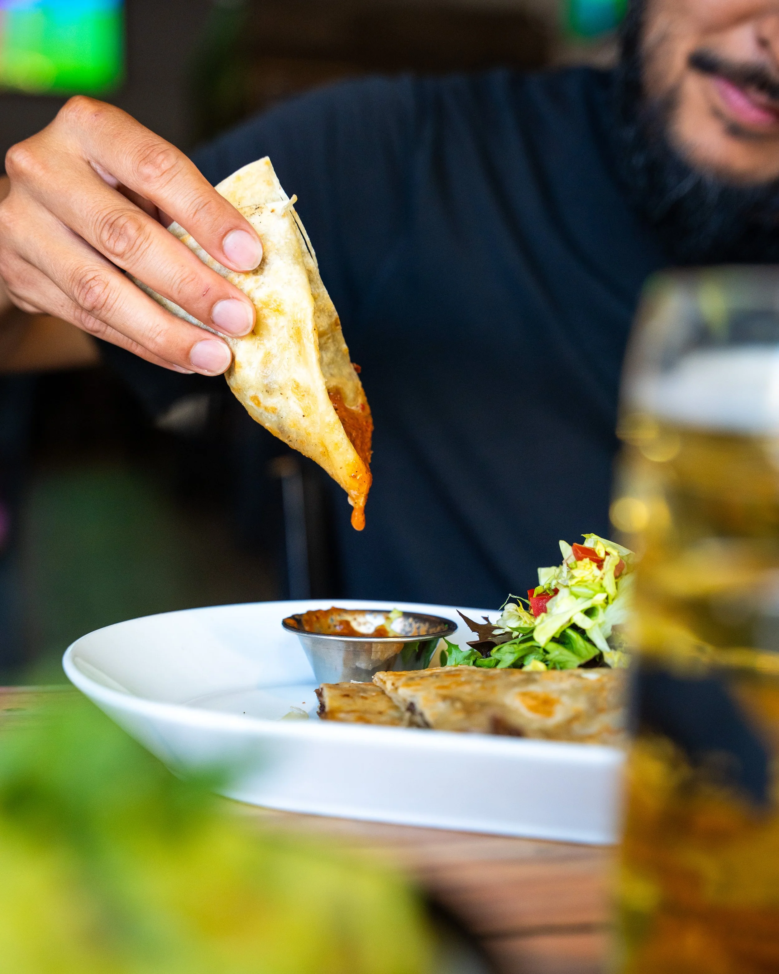 A man wearing a black shirt holding a folded slice of pizza with tomato sauce. On the table, there is a white platter with another slice of pizza, a small salad with tomatoes and greens, and a small container of dipping sauce.