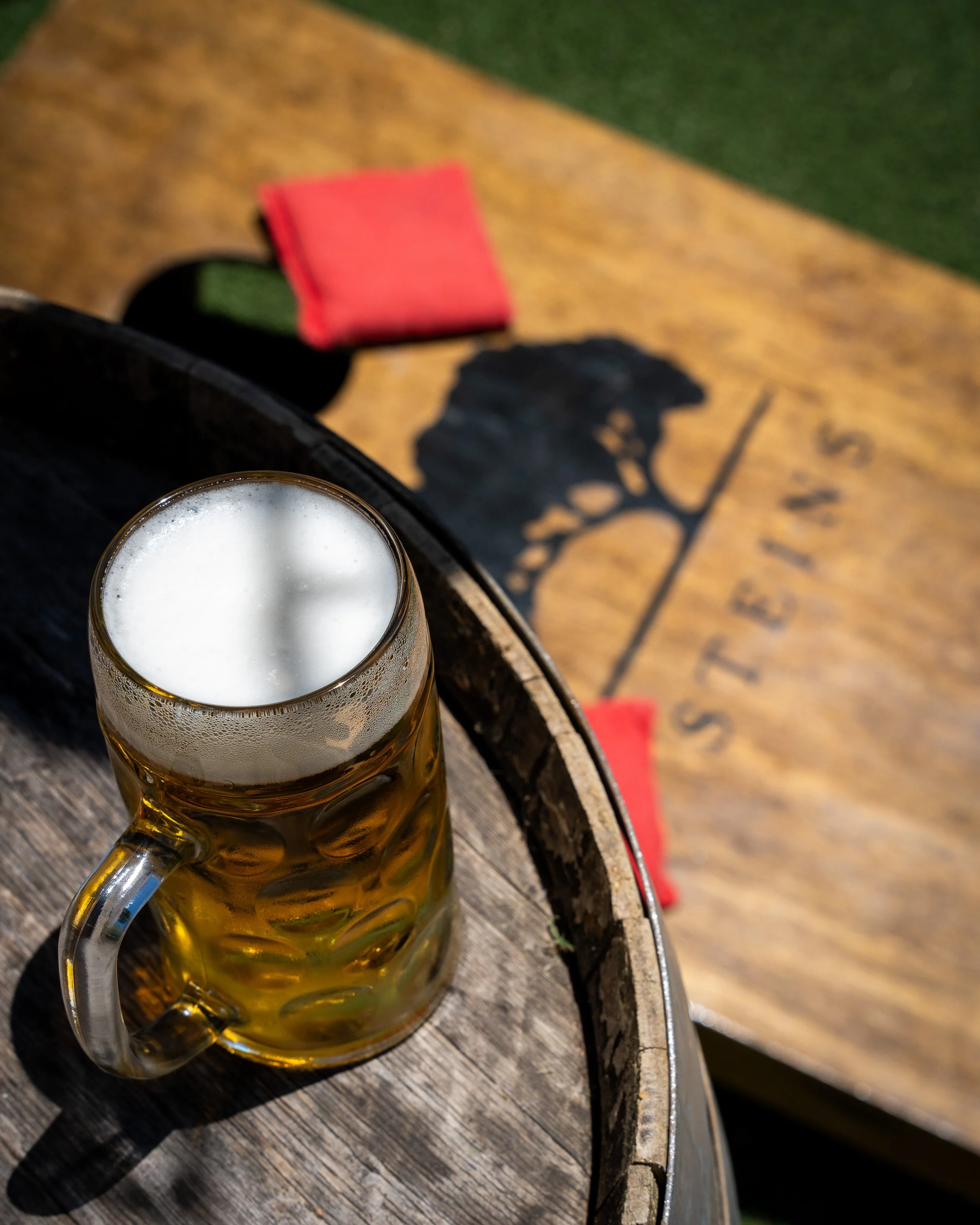 A glass mug of beer with foam on top, placed on a wooden surface, with the shadow of a tree cast onto the surface.