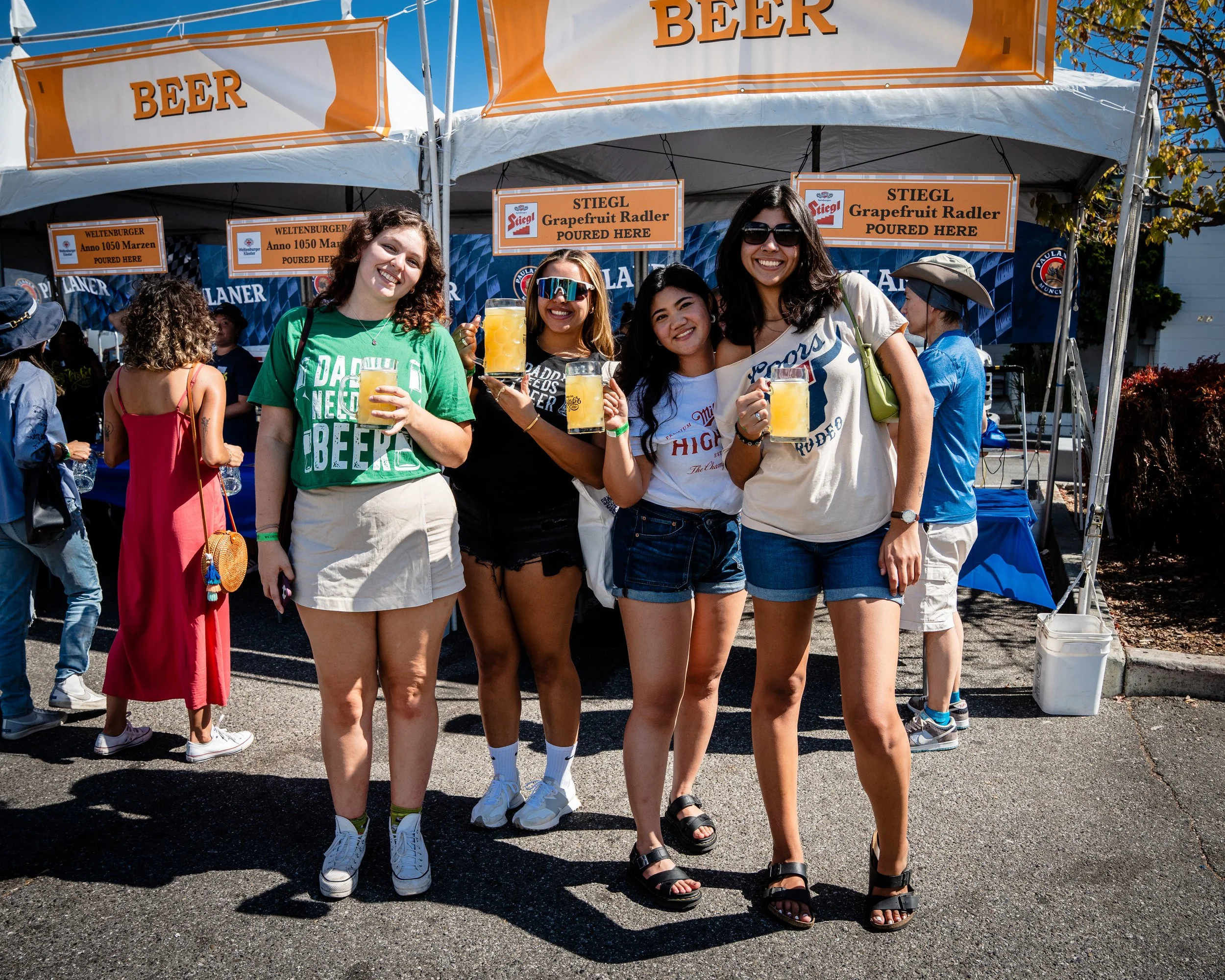 Four women standing outdoors at a beer festival, holding large mugs of beer and smiling. They are in front of a beer stand with a bright orange sign that says 'BEER' and smaller signs advertising grapefruit radler.