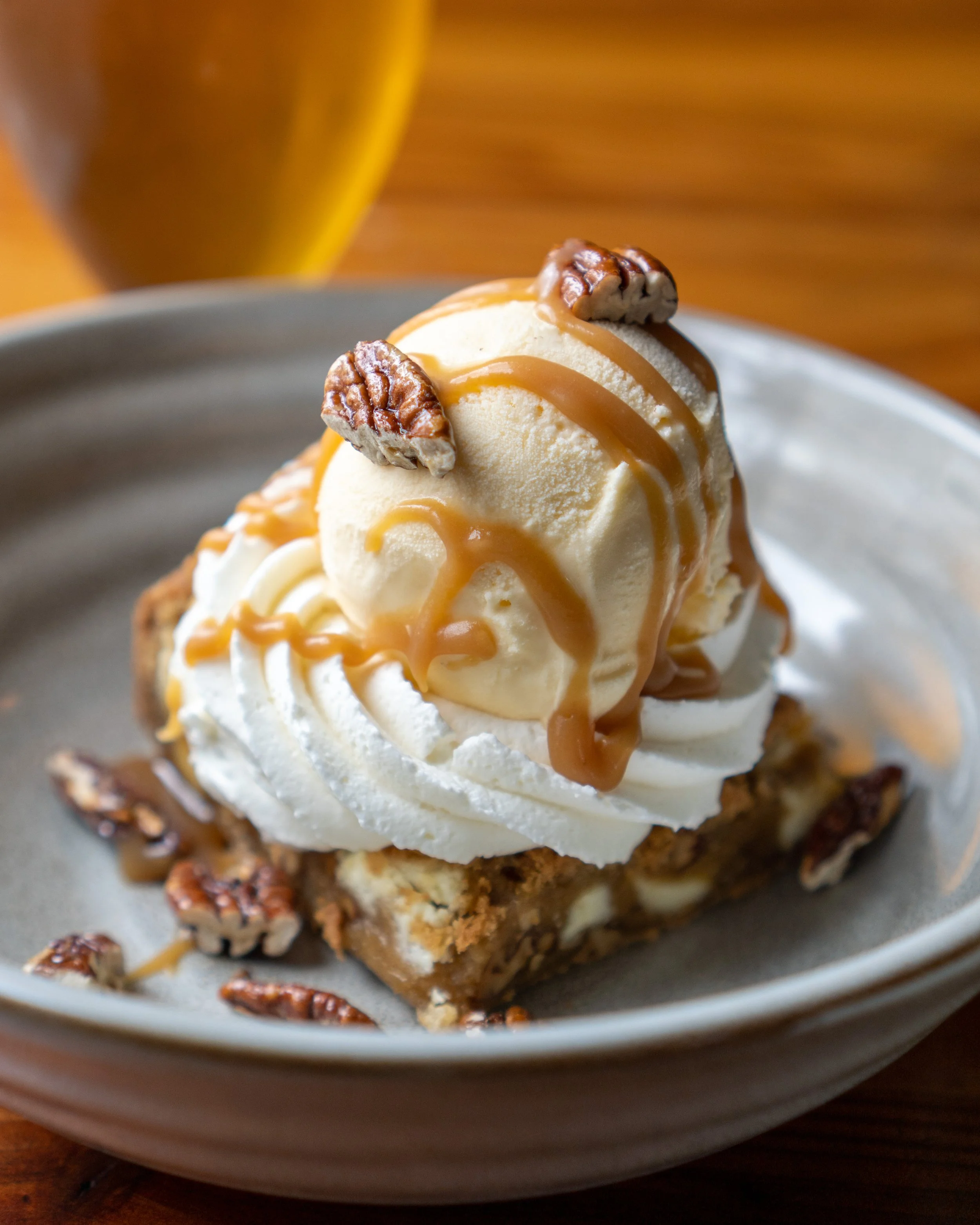 Dessert featuring a scoop of vanilla ice cream on a piece of pecan pie topped with whipped cream, caramel drizzle, and pecans, served on a gray plate.