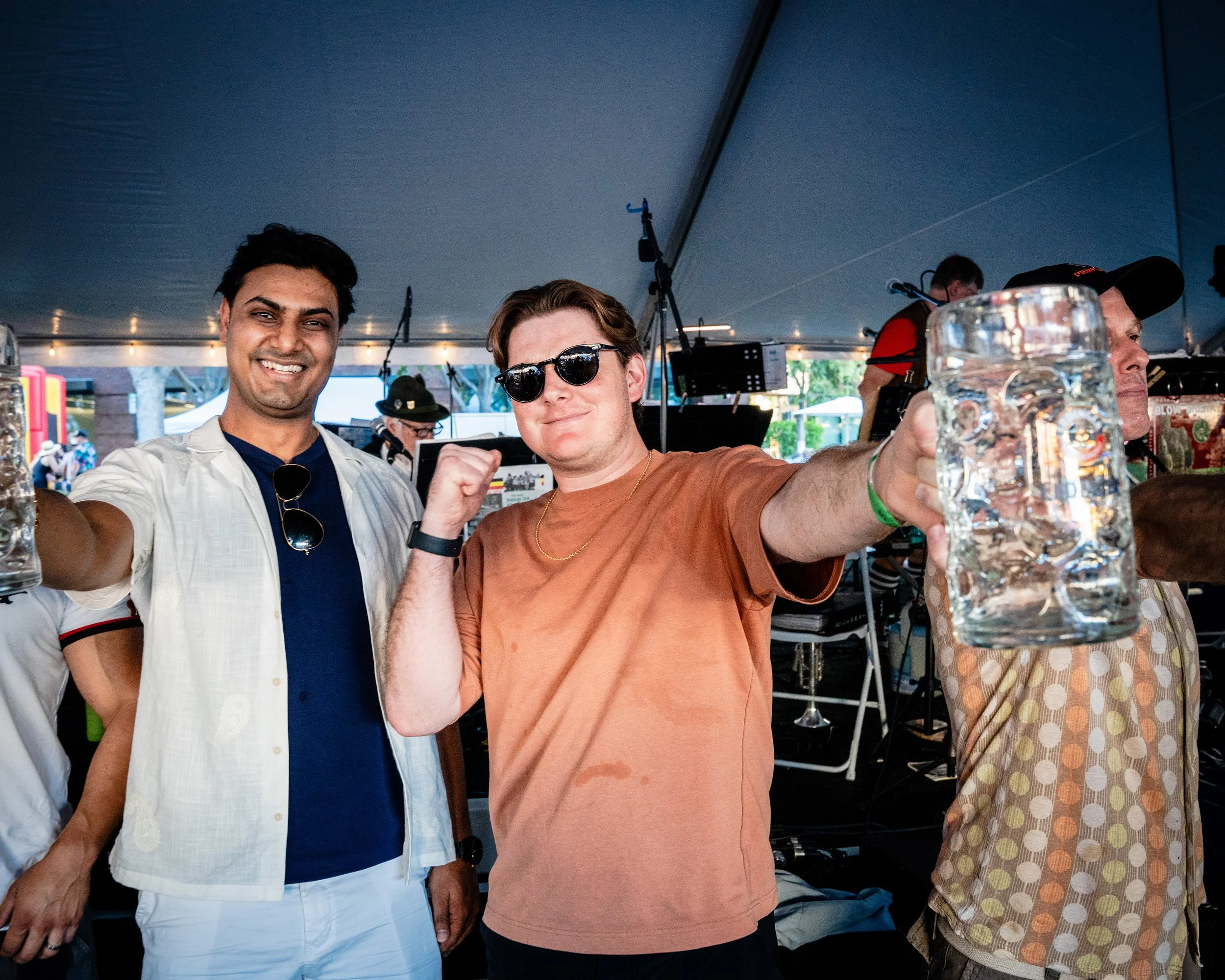 Two men celebrating at an outdoor event, holding up beer mugs, smiling, with a festive tent and other people in the background.