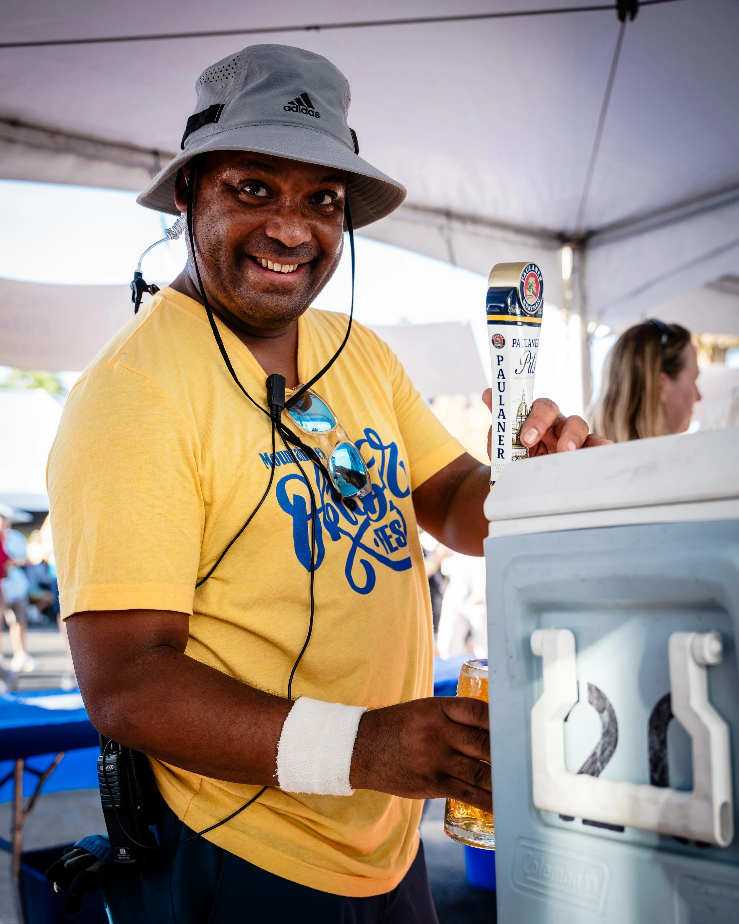Man smiling at a beer stand, wearing a gray hat, yellow t-shirt, sunglasses hanging from his shirt, and a white wristband, pouring a beer from a tap labeled 'Paulaner' at an outdoor event.