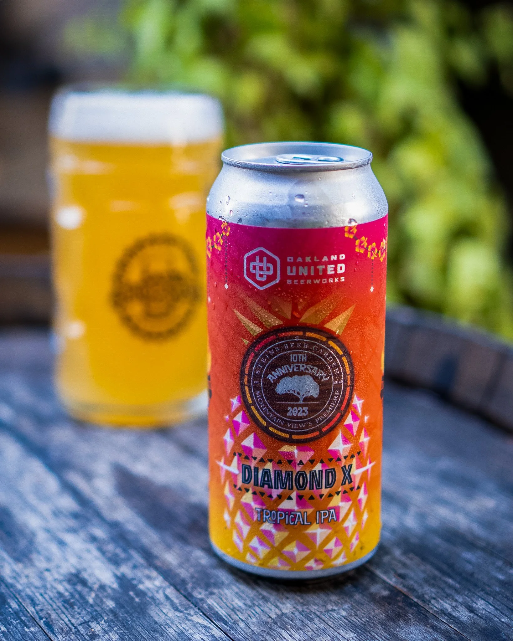 A can of Diamond X Tropical IPA beer from Oakland United Beerworks in front of a glass of beer on a wooden table with greenery in the background.