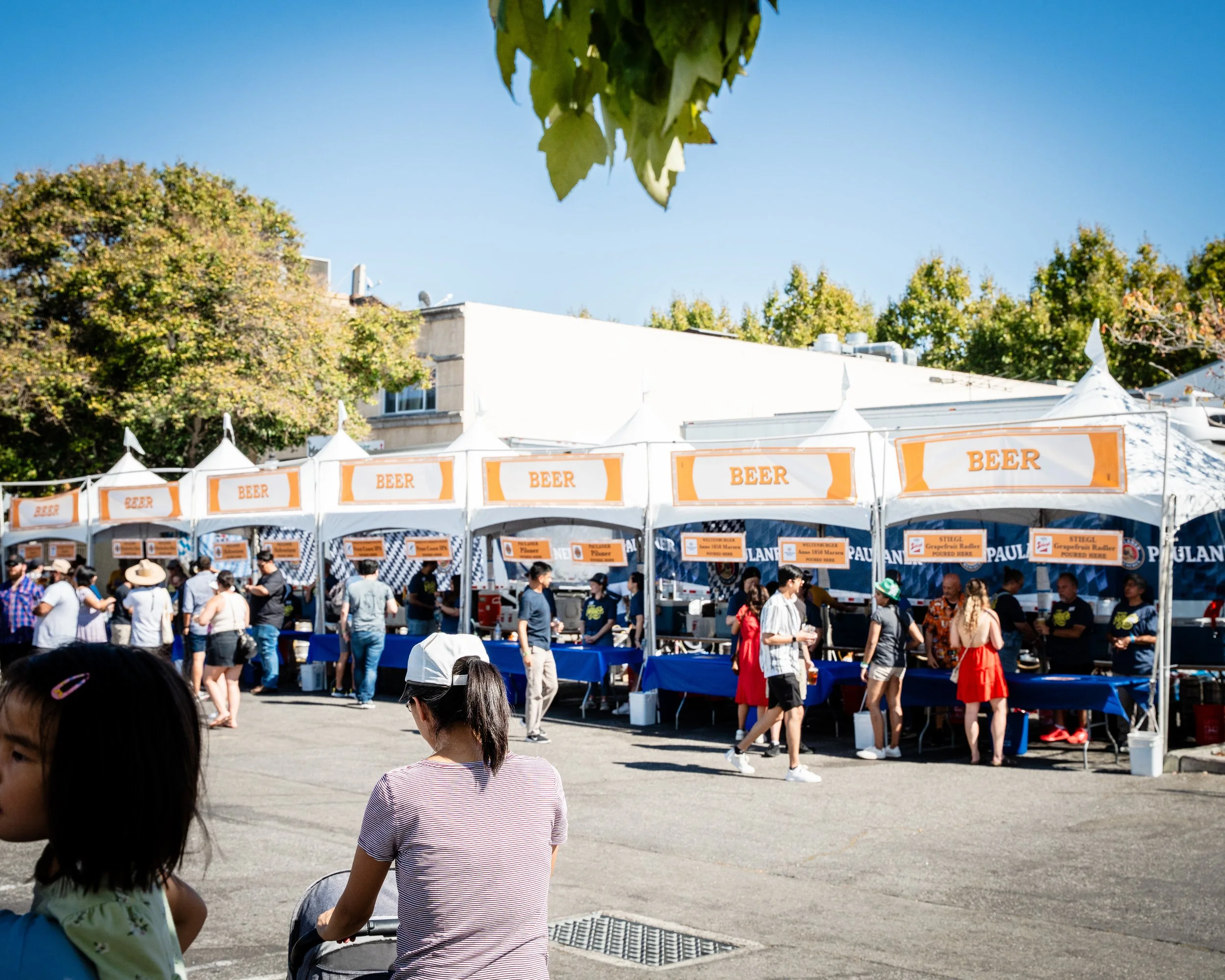 A busy outdoor festival with white tents labeled 'BEER' and signs for various food vendors, with people walking, talking, and sitting on the street on a sunny day at festival.