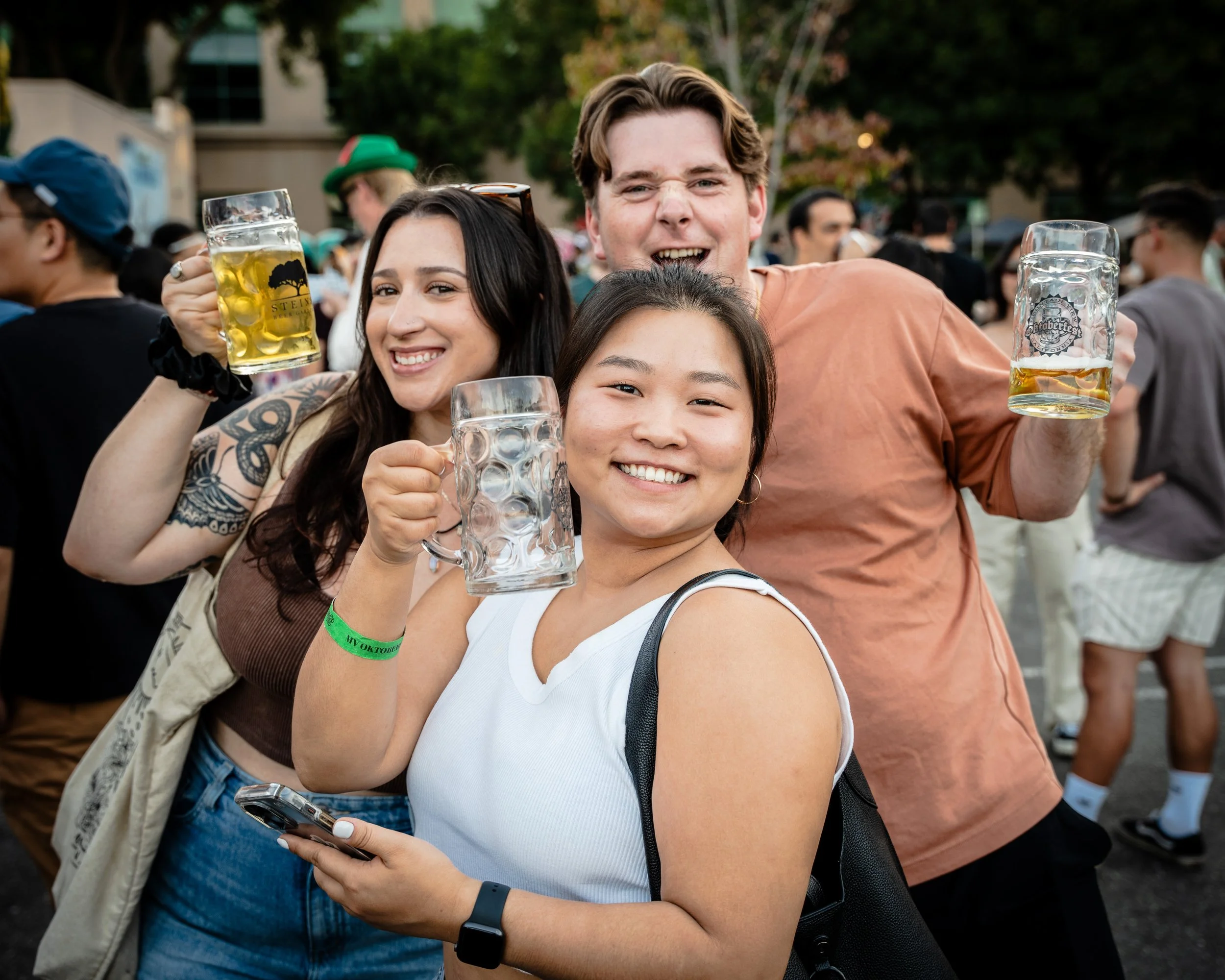 Three friends at a social event outdoors, holding large beer mugs and smiling at the camera at Oktoberfest.
