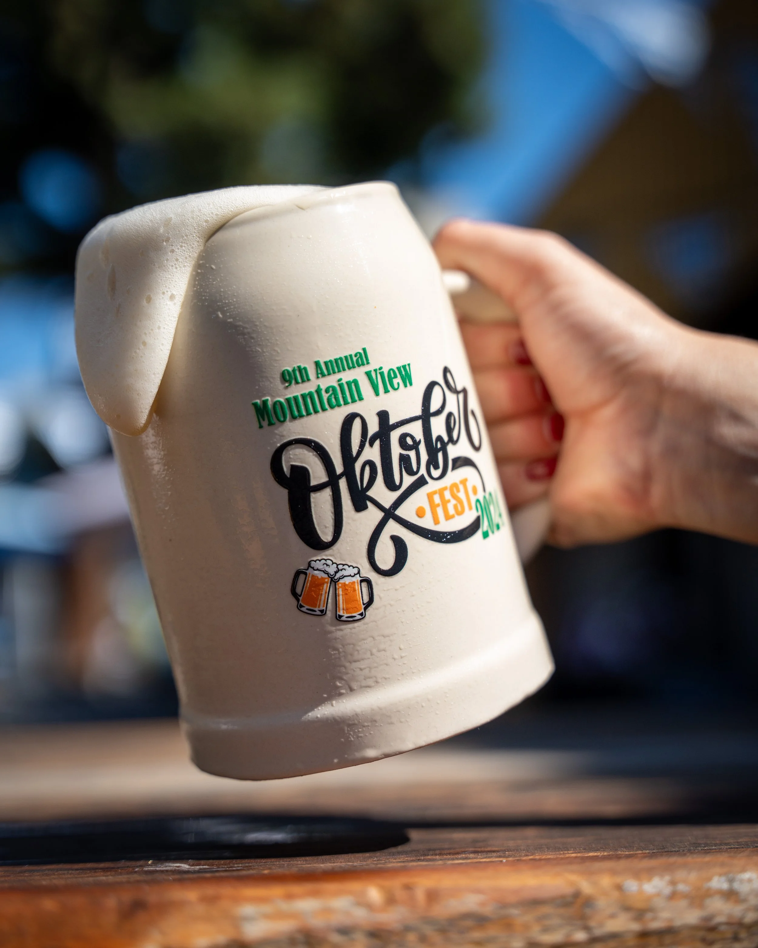 A hand holding a large beer stein with foam overflowing, featuring a logo for the 9th Annual Mountain View Oktoberfest, with a background of outdoor festival scene.