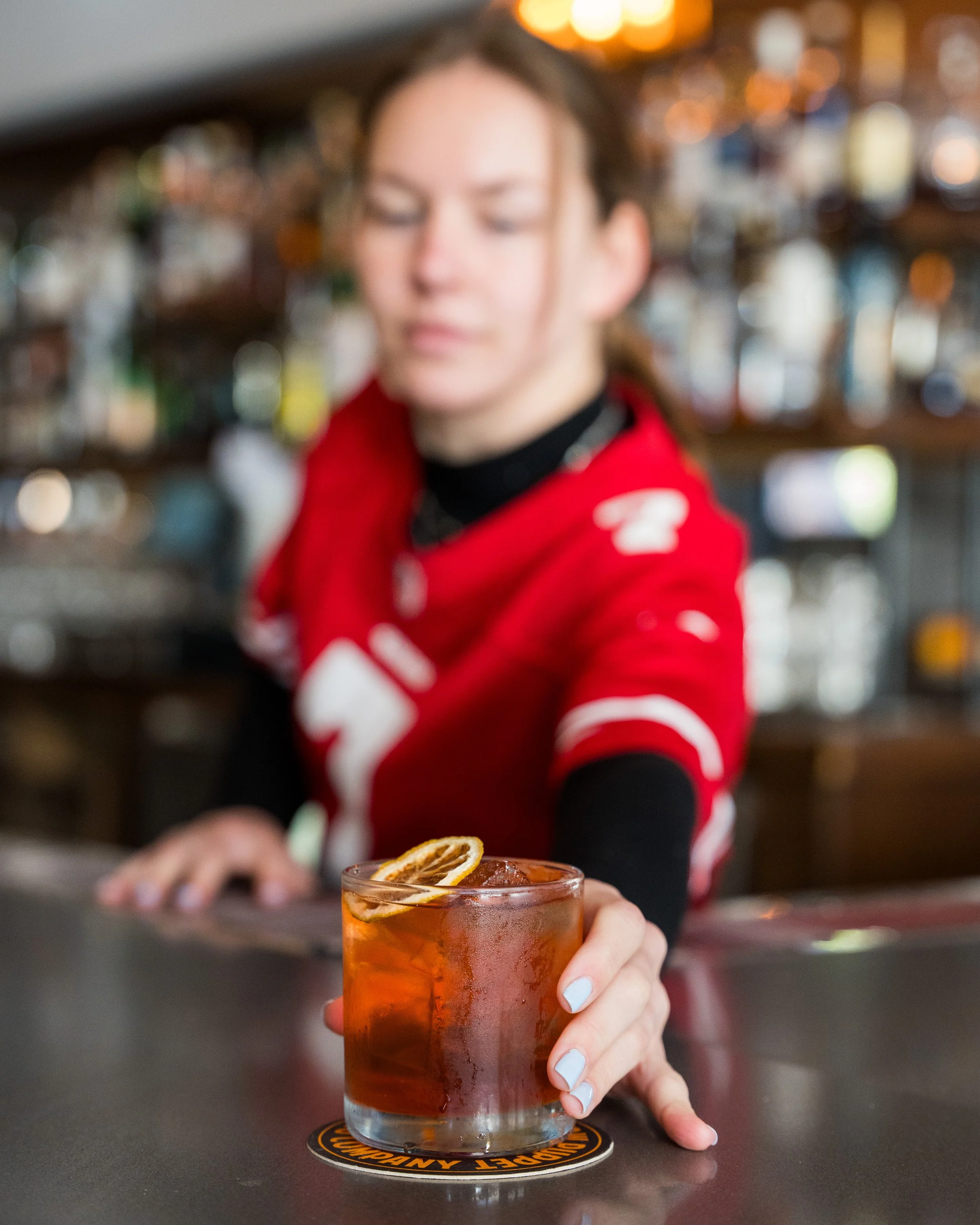 A bartender reaching across a bar counter presenting a glass of iced cocktail garnished with a lemon slice.