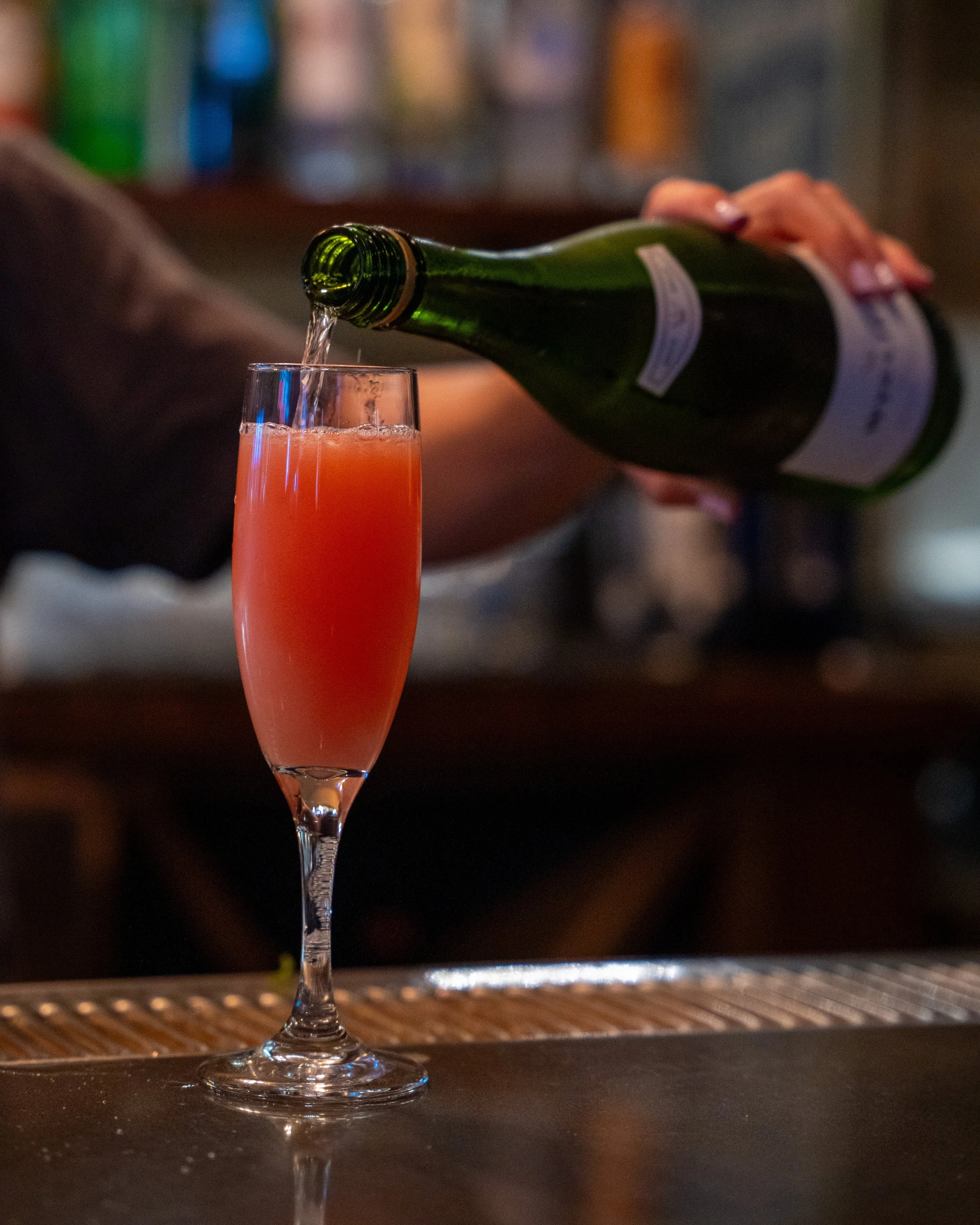 A person is pouring a pink cocktail from a green bottle into a tall champagne flute on a bar counter.