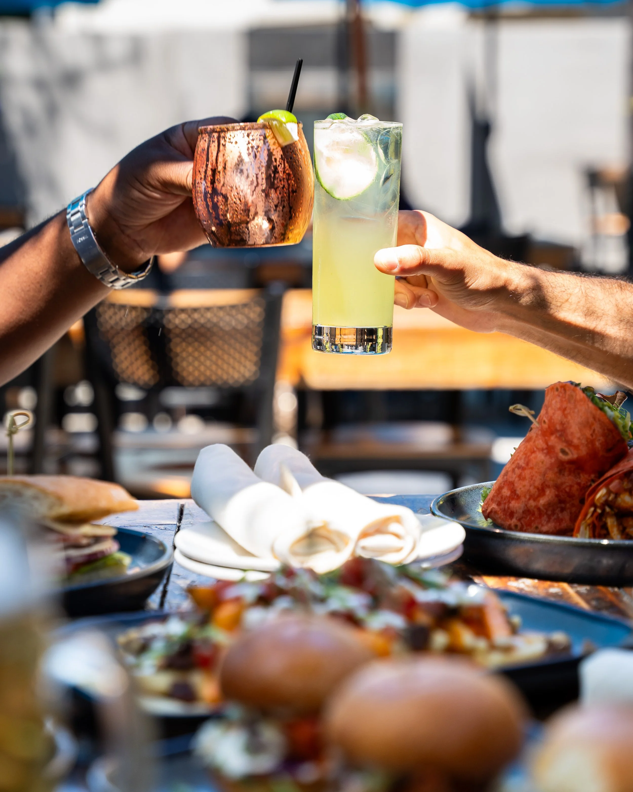 Two people clinking drinks, one with a copper-colored cocktail and the other with a light green beverage, at an outdoor dining table with food including tacos and salads.