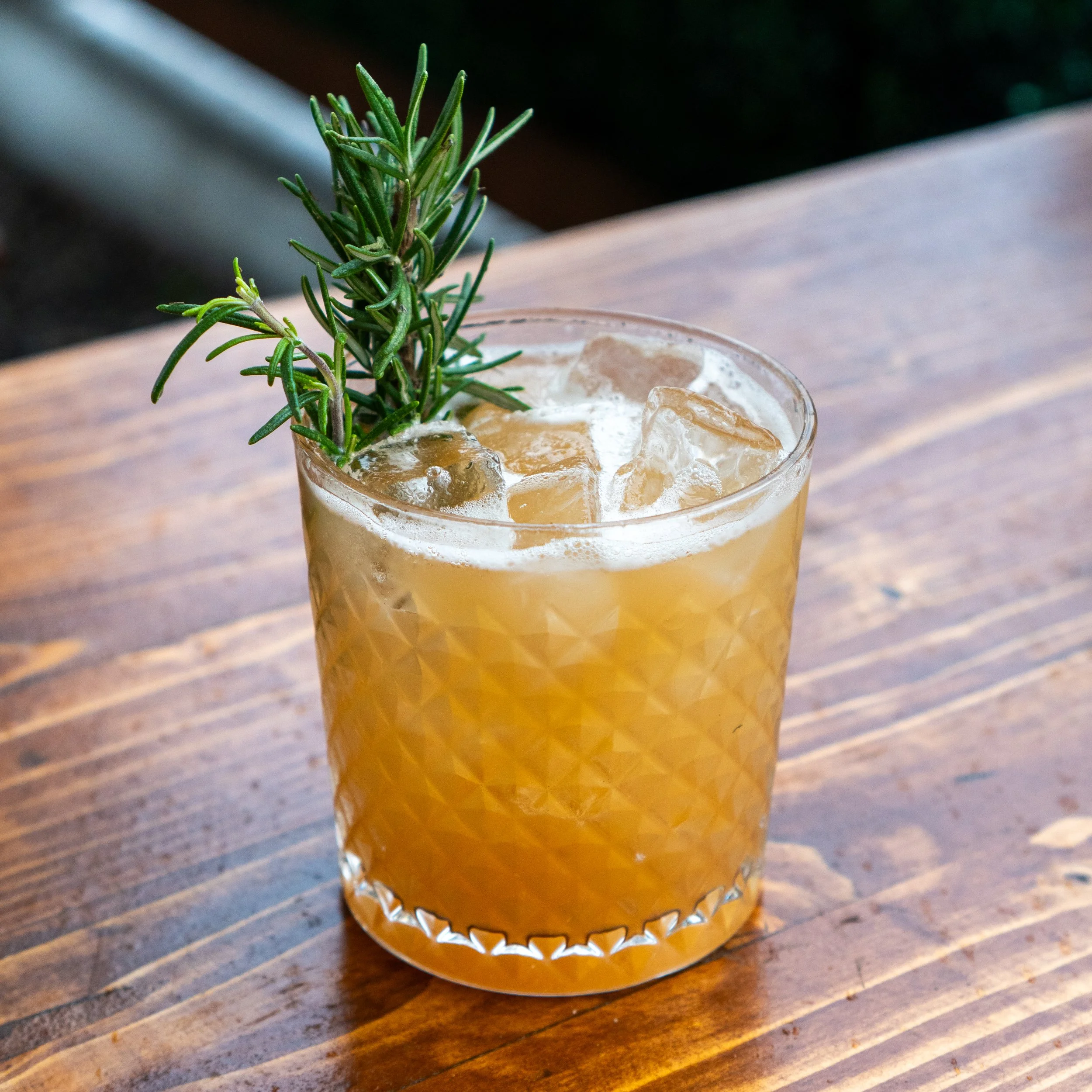 A glass with a textured pattern containing a yellowish cocktail with ice and a sprig of rosemary, placed on a wooden table.