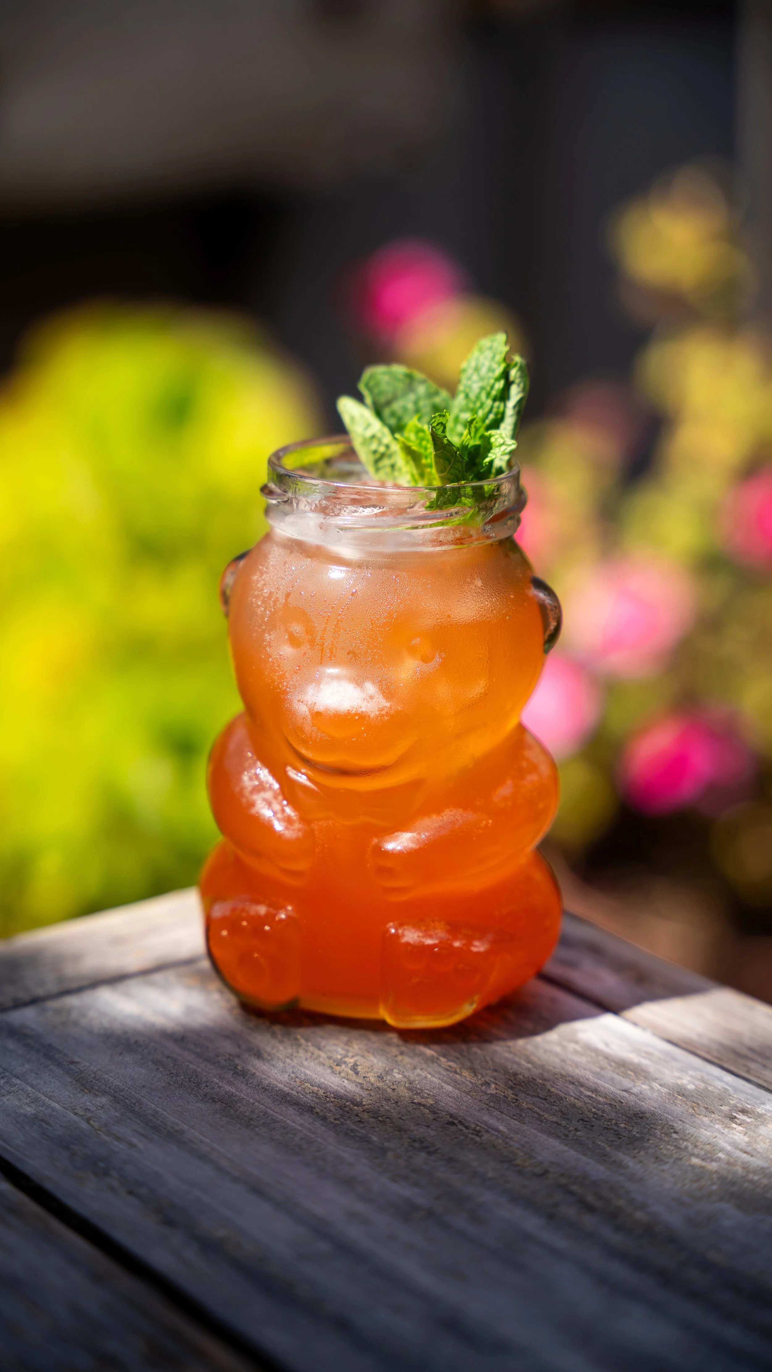 An orange gummy bear-shaped glass filled with an orange-colored beverage topped with fresh mint leaves, placed on a wooden surface outdoors with blurred colorful flowers in the background.