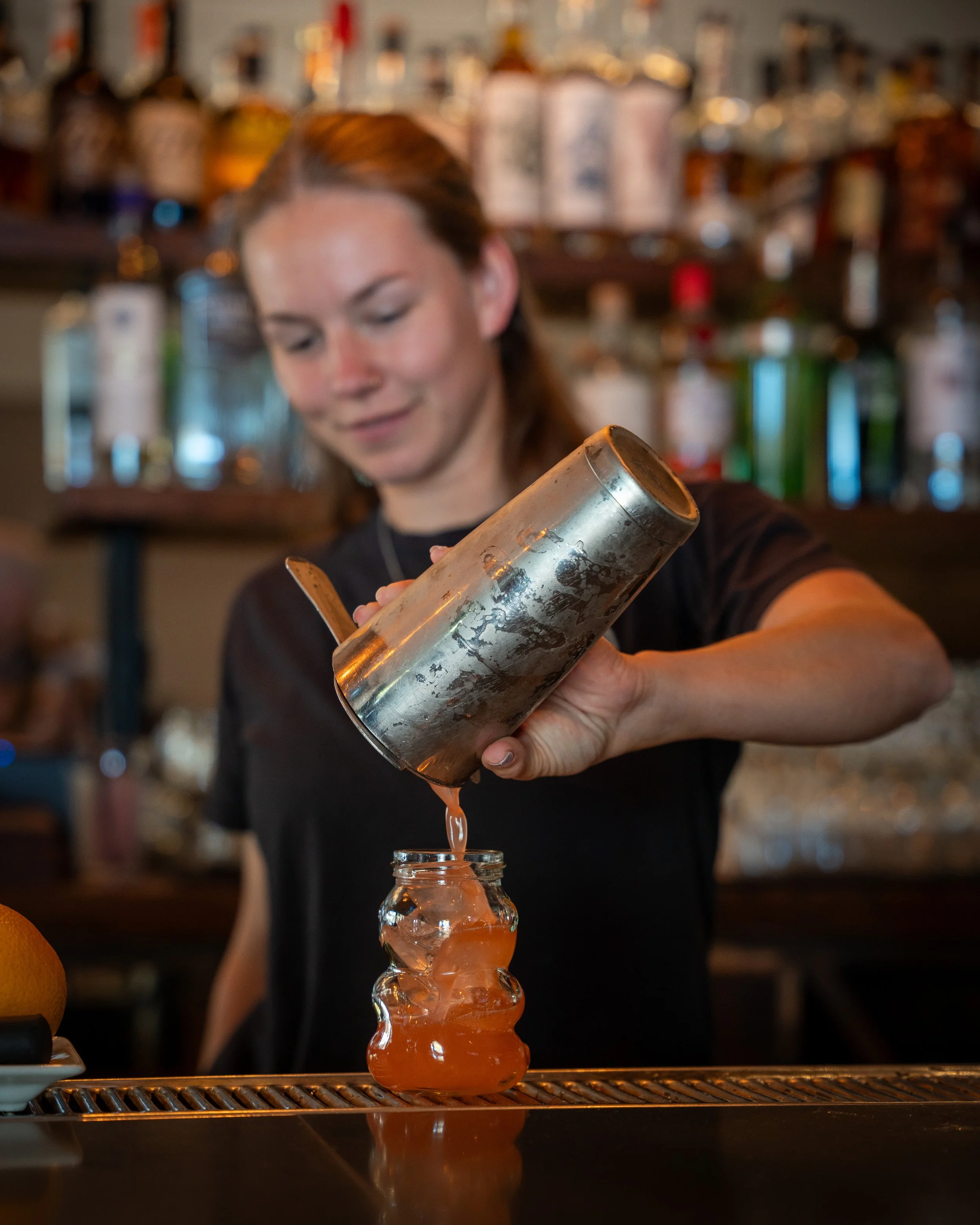 A bartender pouring a cocktail from a shaker into a glass in a bar setting.