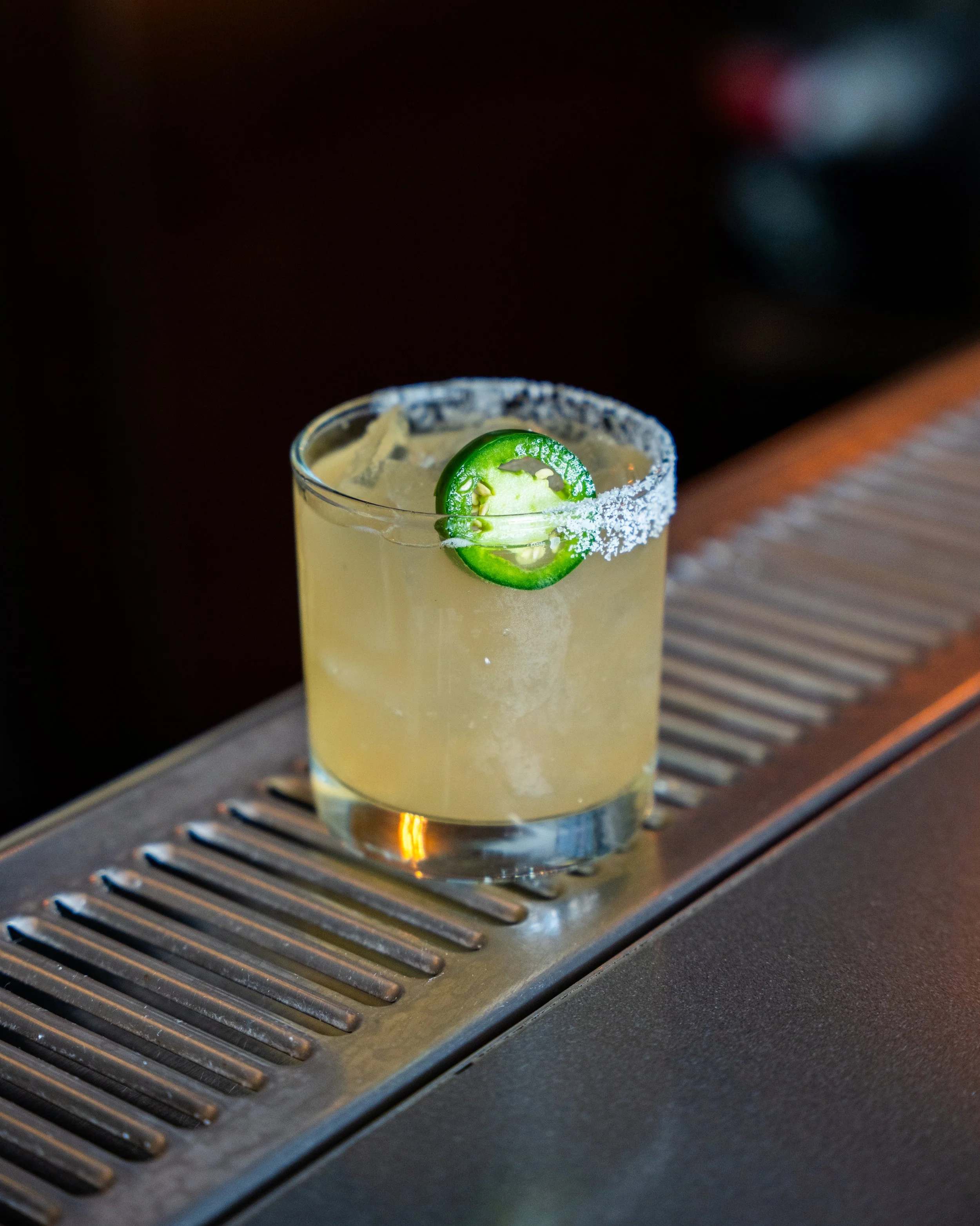 A cocktail in a short glass with a lime slice and salt rim on a bar counter.