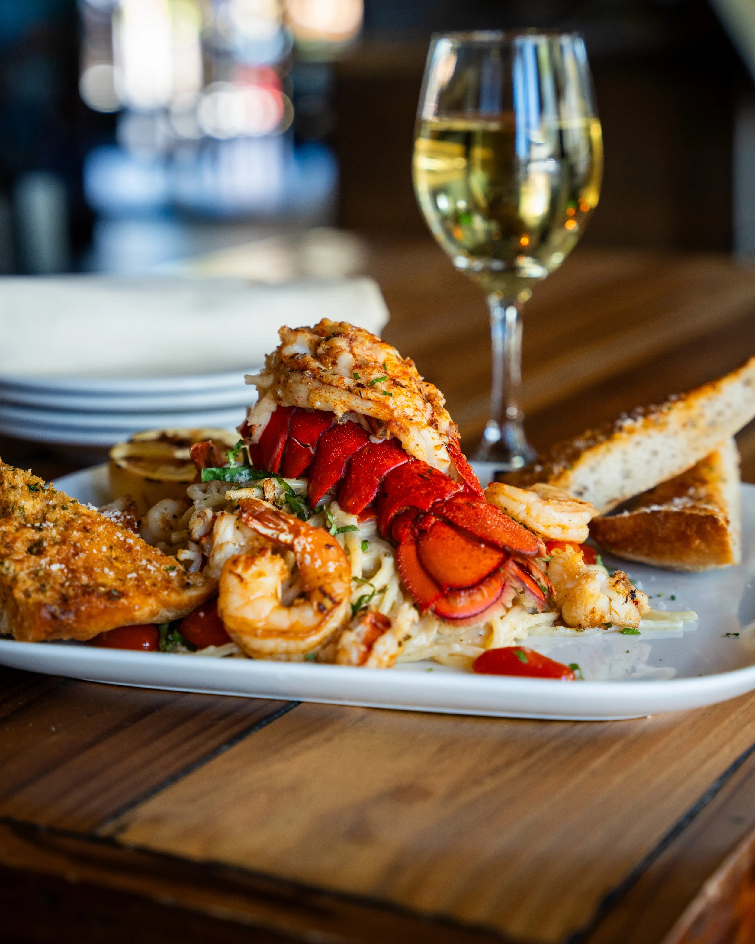 Lobster tail with shrimp, pasta, garlic bread, and a glass of white wine on a wooden table in DC restuarant.