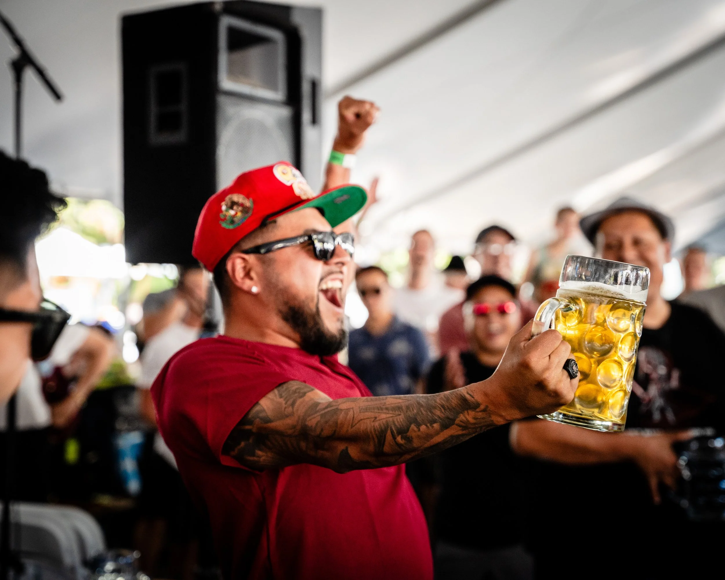 A man wearing sunglasses, a red cap with patches, and a red shirt, holding a large glass mug of beer, laughing and celebrating at a crowded event under a tent.