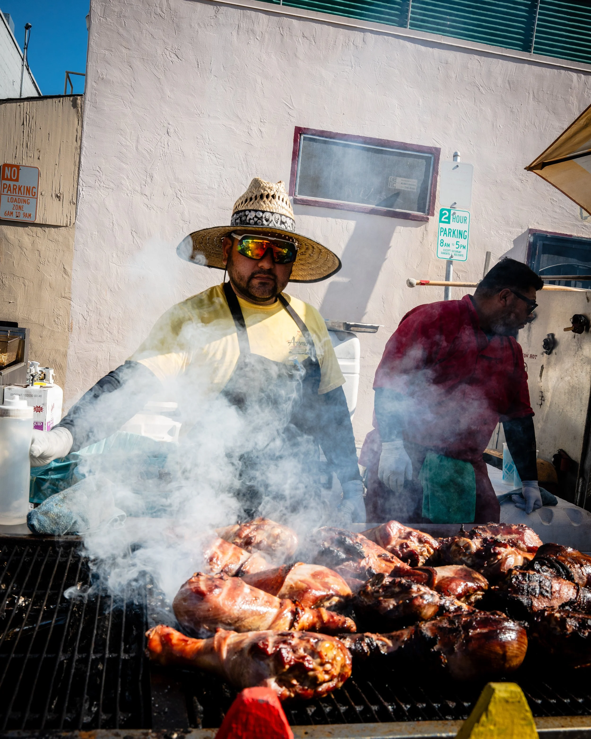 Two men grilling large pieces of meat outdoors. The man on the left wears a large straw hat, sunglasses, a yellow shirt, and a black apron, standing behind a grill with smoke rising from the meat. The man on the right is dressed in a red shirt and ap