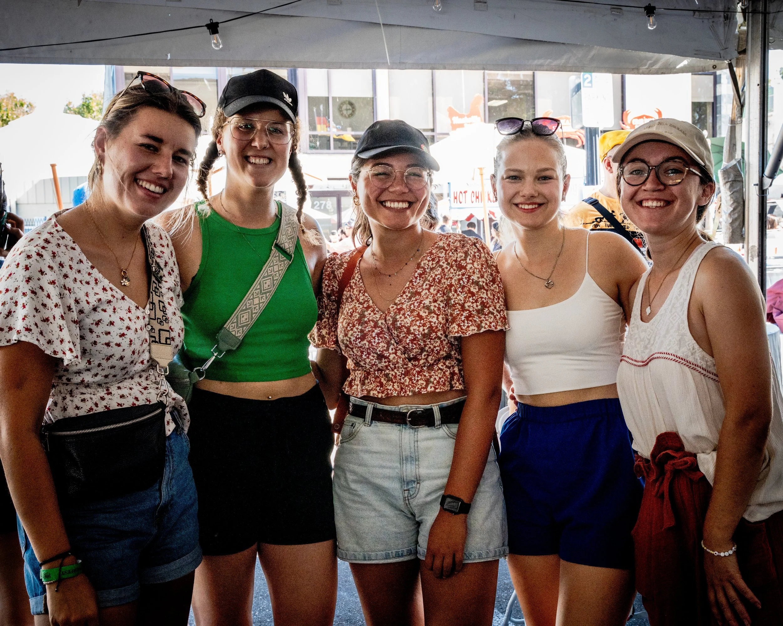 Group of five young women smiling, standing together at an outdoor event, dressed casually with summer clothing and accessories.