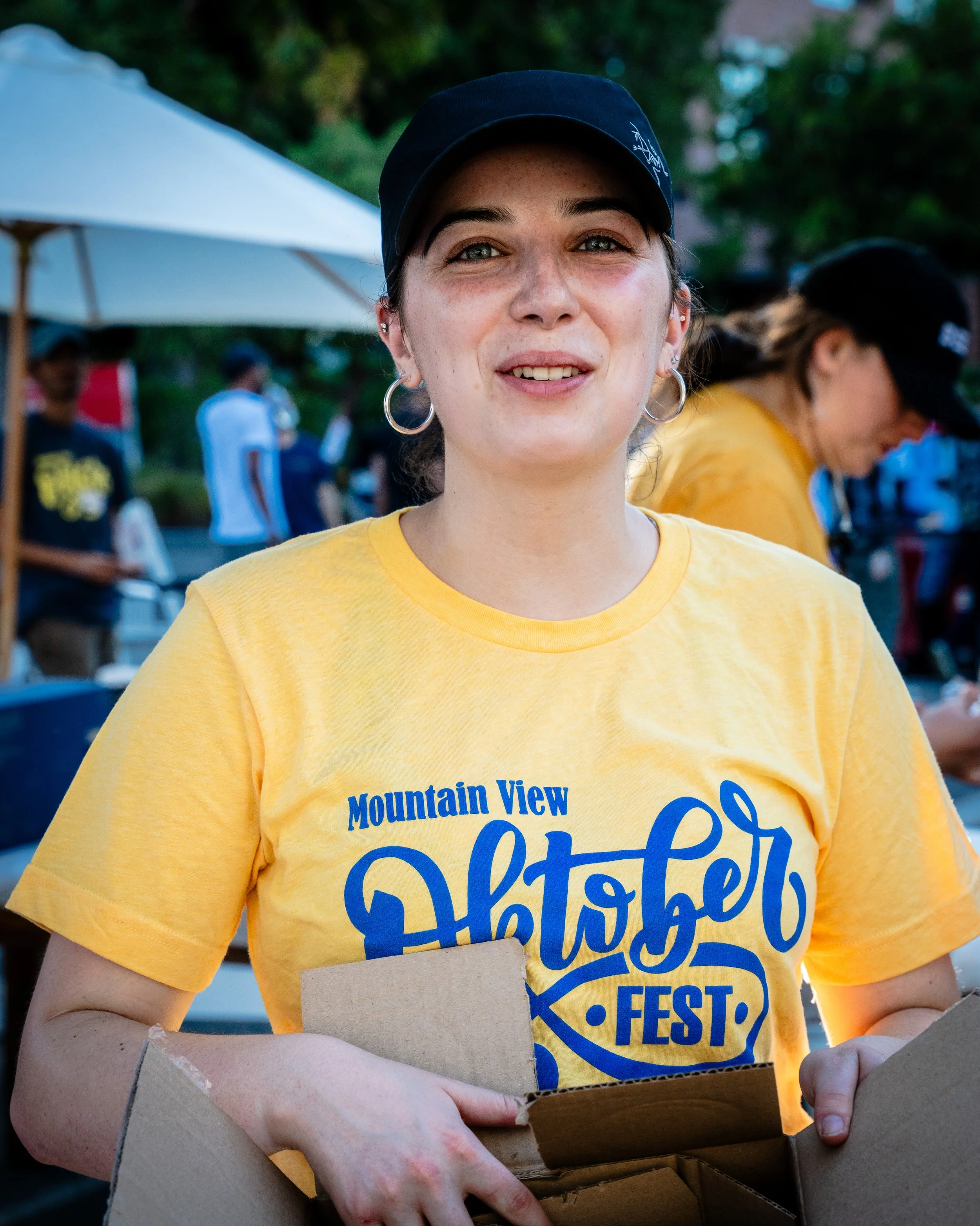 Young woman with earrings and a black hat smiling at an outdoor festival, wearing a yellow t-shirt that says 'Mountain View Dhotar FEST' and holding an open cardboard box.