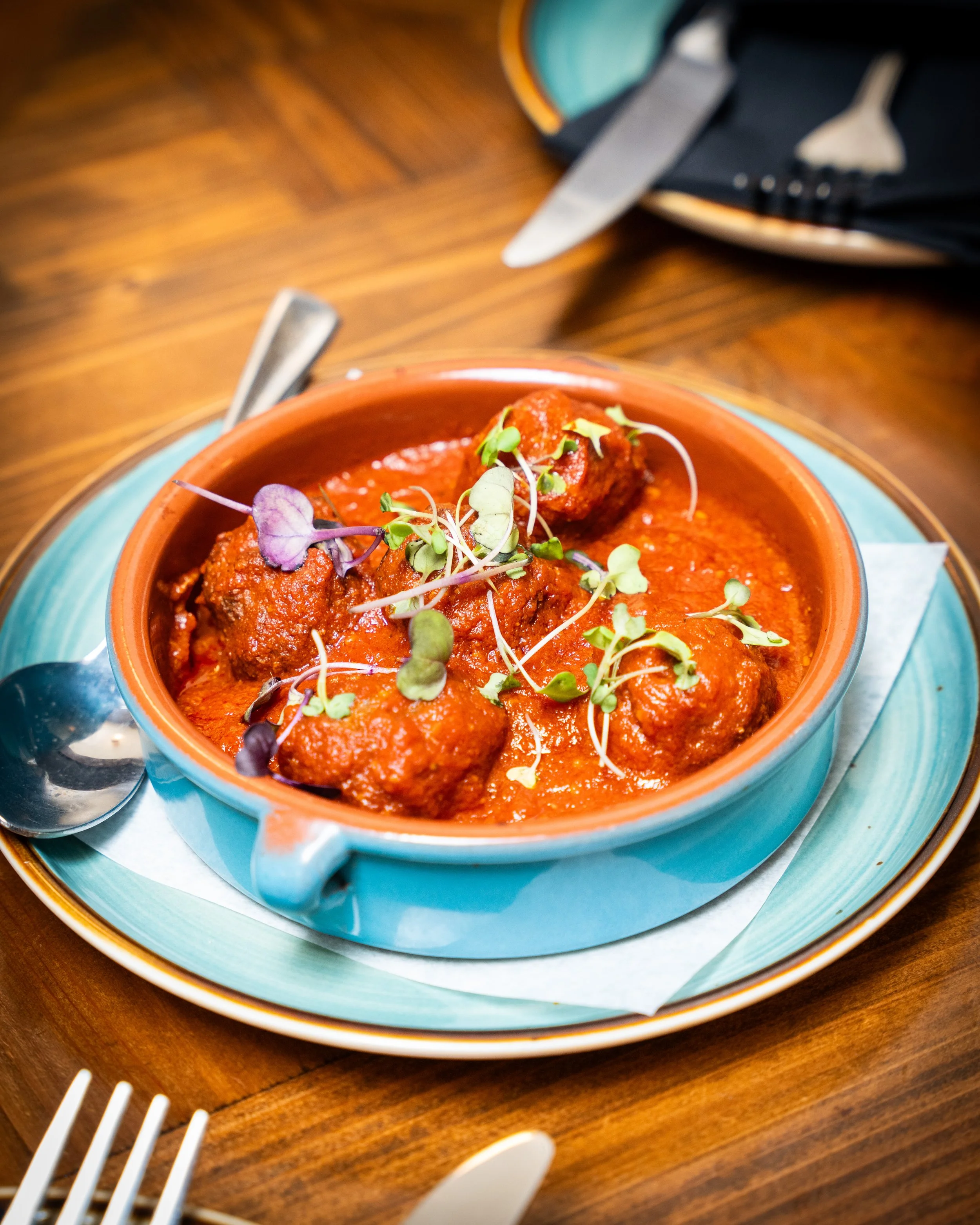 A bowl of meatballs in tomato sauce garnished with microgreens, served on a blue plate with a spoon and utensils nearby.