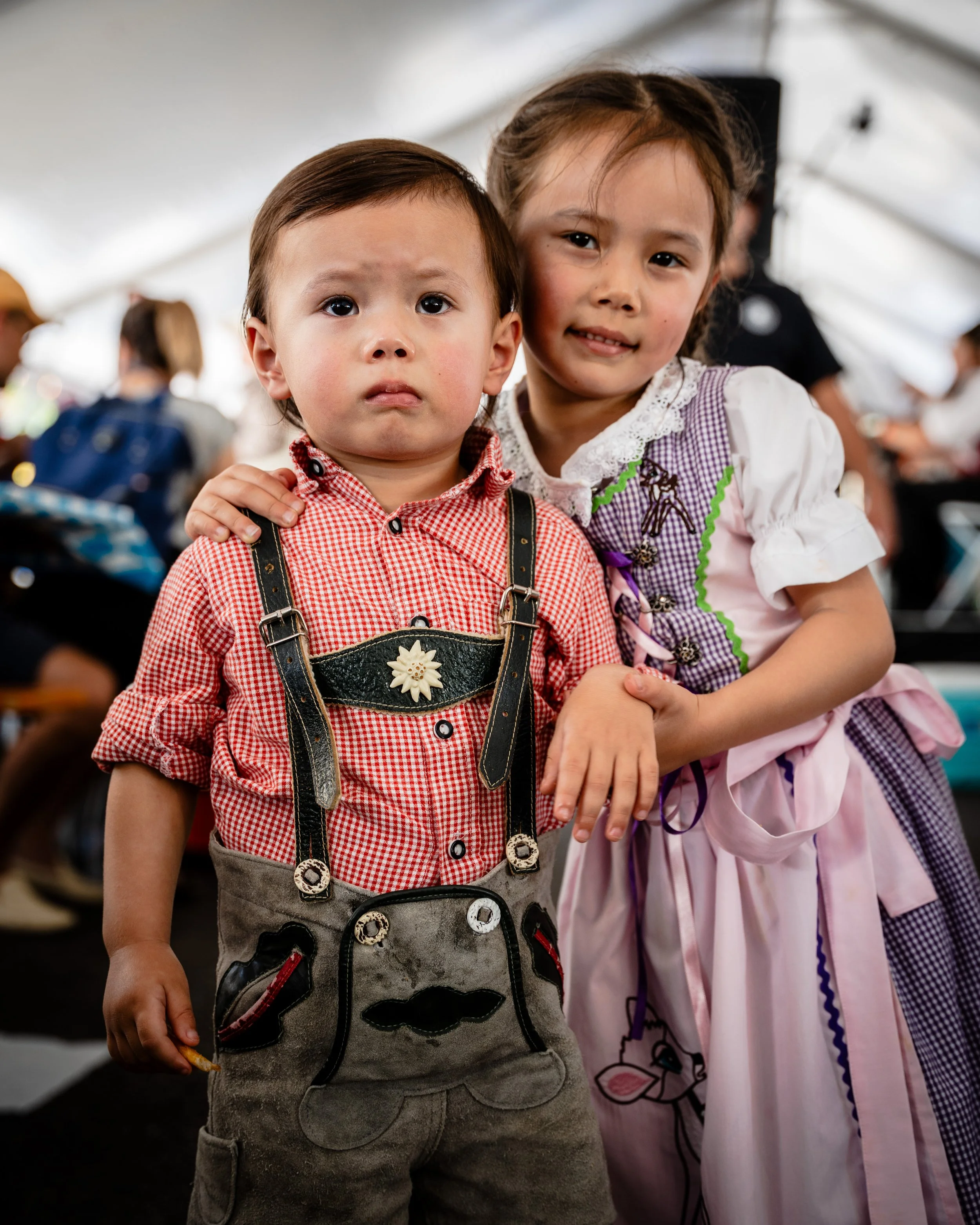 A young boy and girl in traditional German attire at a festive event inside a large tent. The boy wears lederhosen and a red checkered shirt, looking serious; the girl in a colorful dirndl dress, smiling and holding the boy's hand.