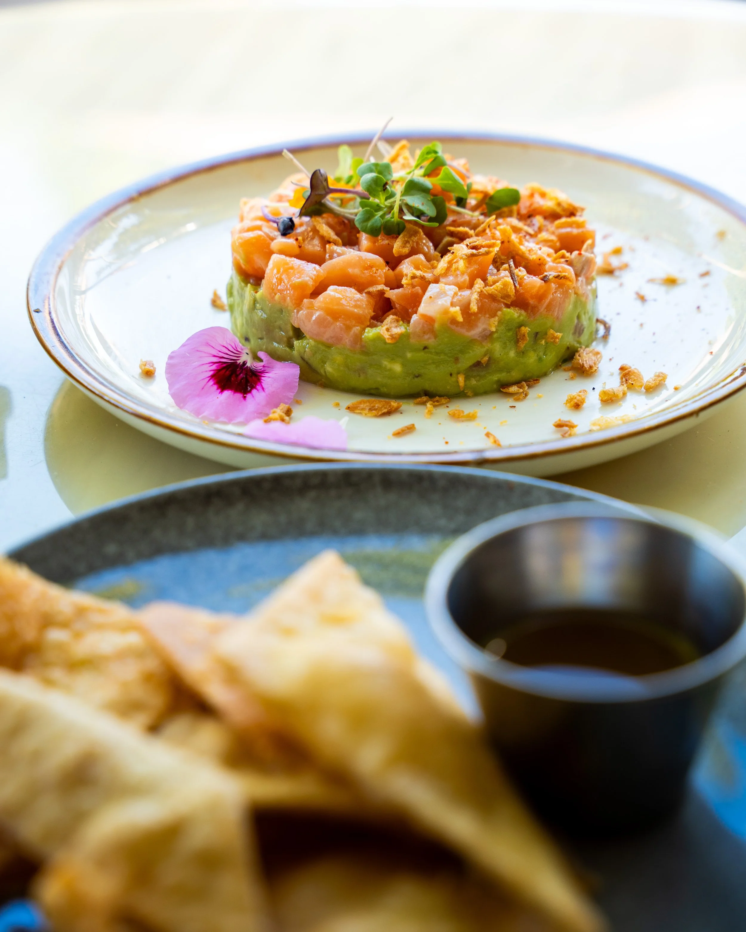 A plate of salmon tartare topped with microgreens, garnished with a pink edible flower, served with avocado, crispy onions, and a side of soy sauce.