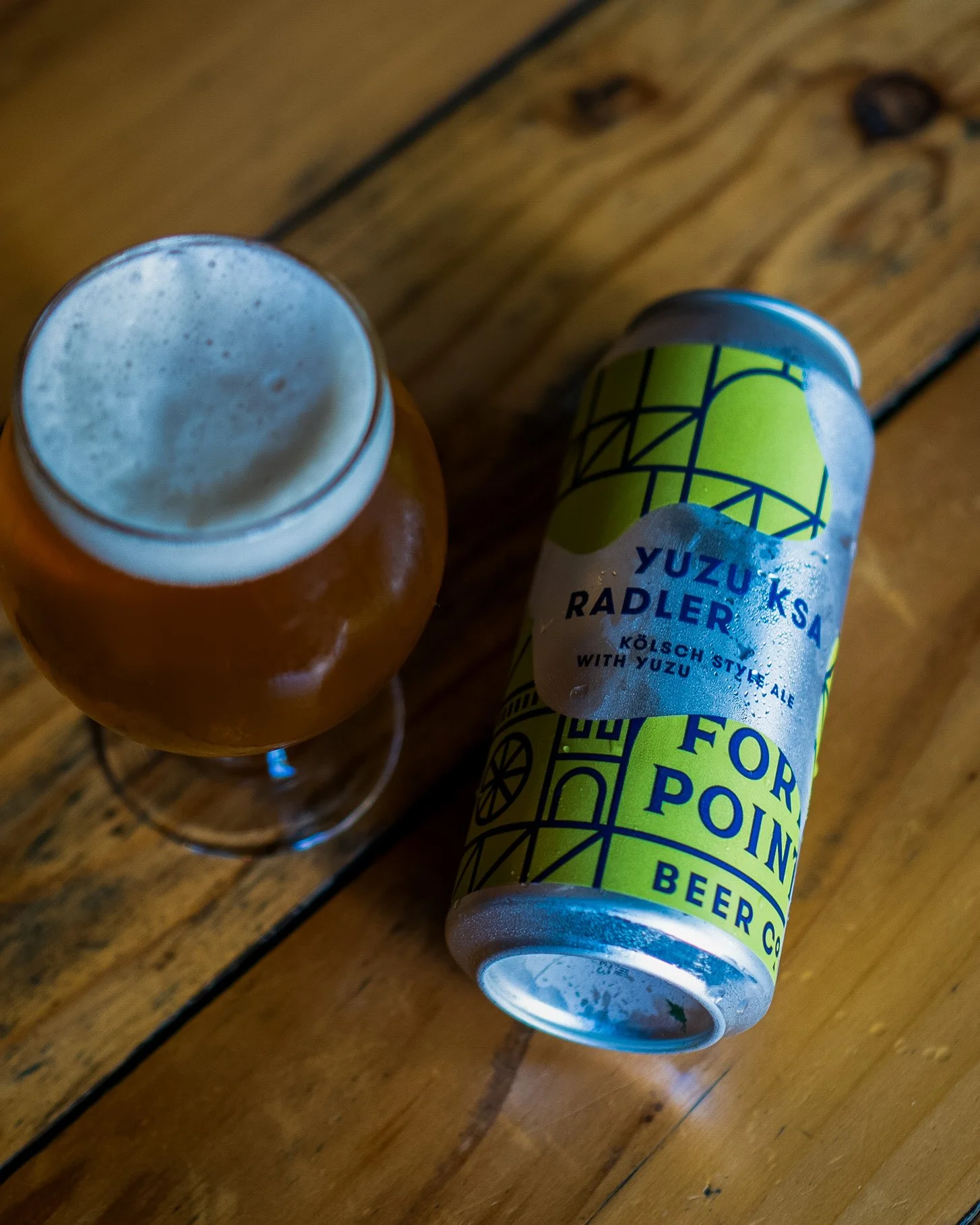 A can of Yuzu Kosa Radler beer with a yellow and green label lying on a wooden table next to a glass of beer with foam on top.