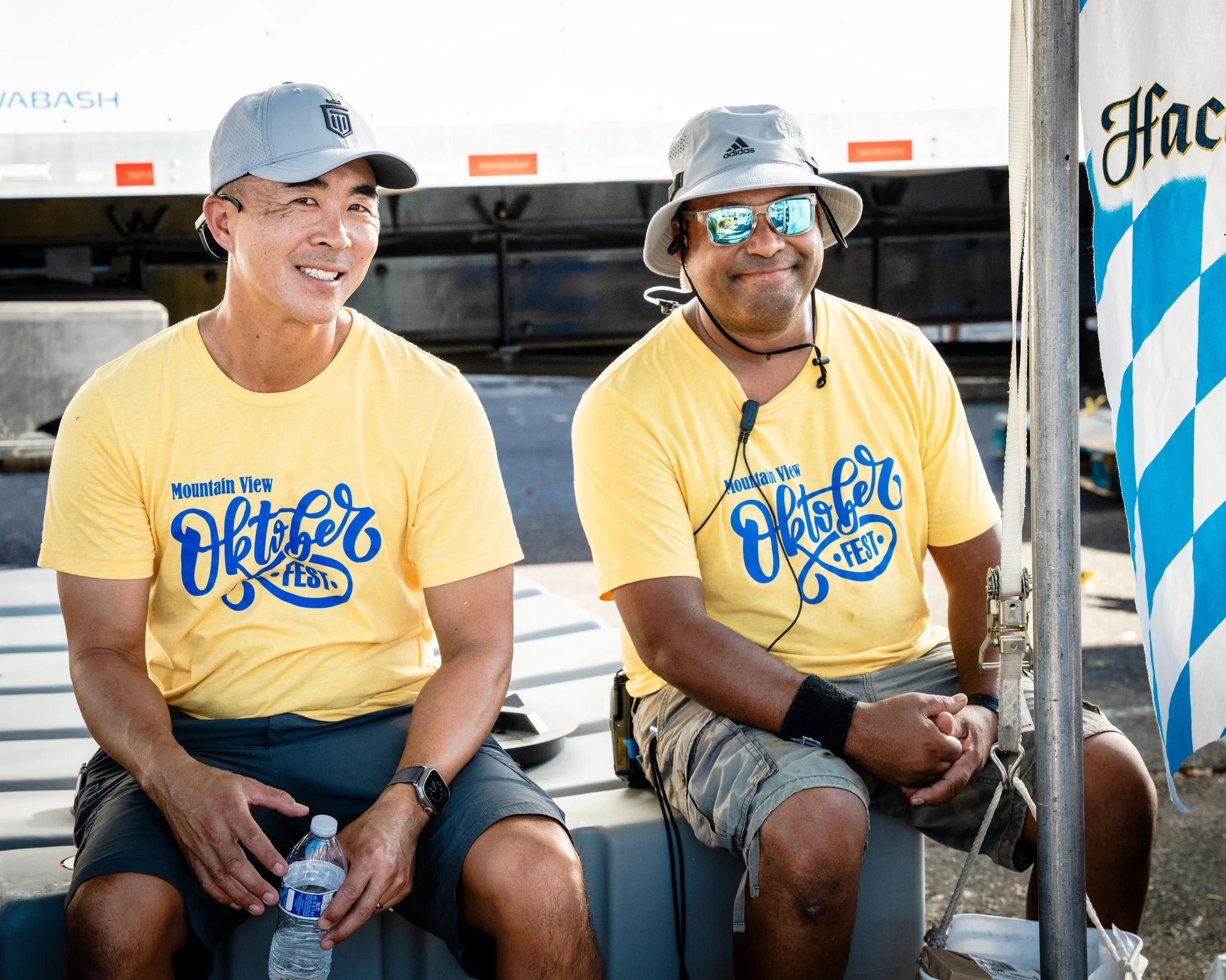 Two men wearing yellow t-shirts with blue text sitting outdoors at the Oktoberfest event. They are smiling, with one holding a water bottle, and the other wearing sunglasses and a bucket hat. There is a Bavarian flag and some flags visible behind the