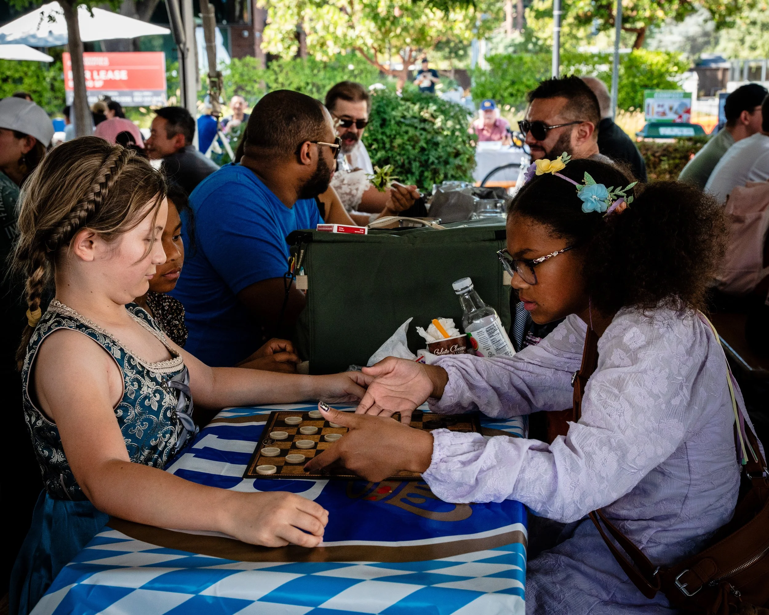 Two girls playing checkers at an outdoor table, one girl with braided hair and the other with floral headband, surrounded by people at a lively outdoor event.