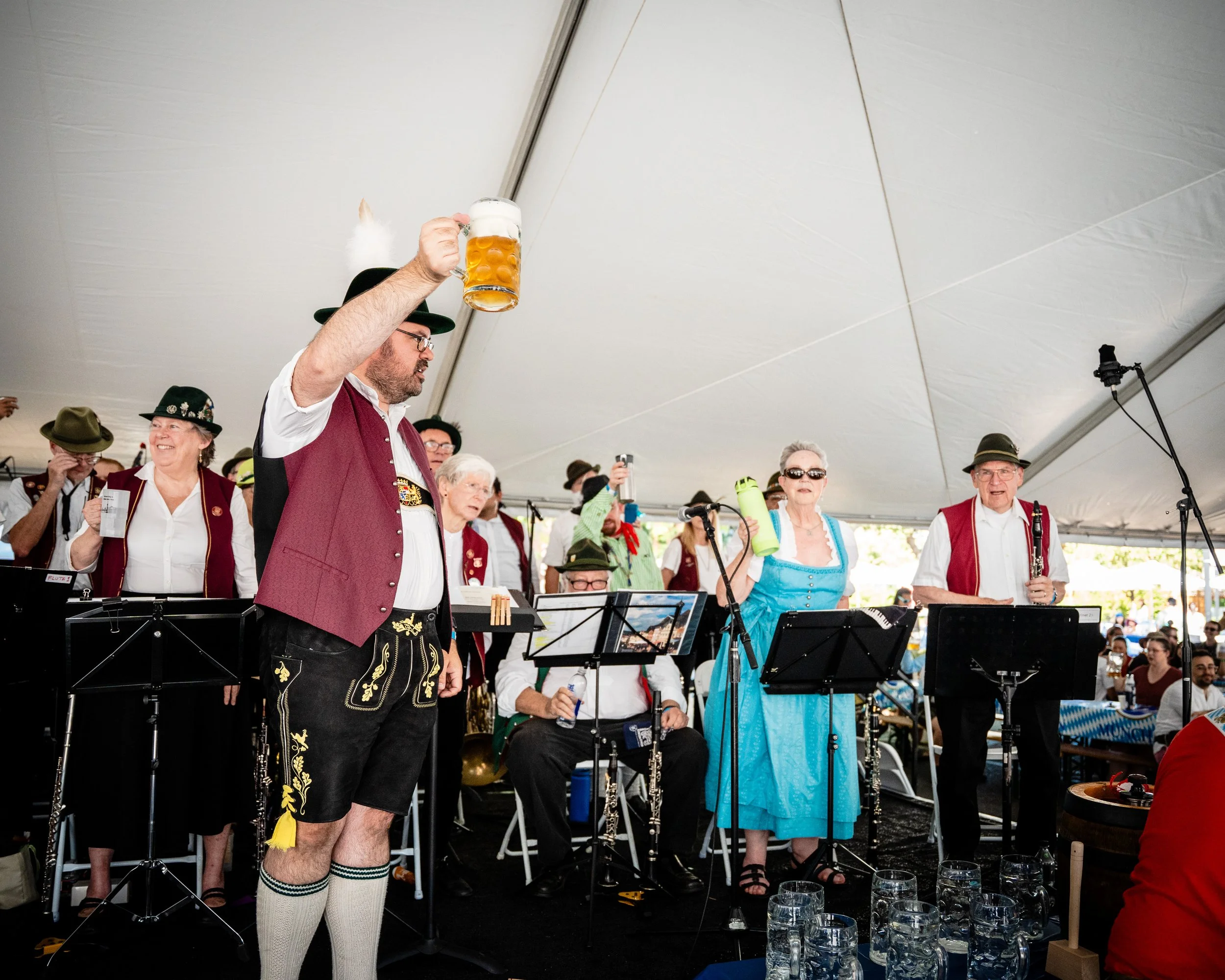 A man in traditional Bavarian attire holding a beer mug at a festive gathering under a large tent, with other people, some also in traditional clothing, playing musical instruments and wearing hats.