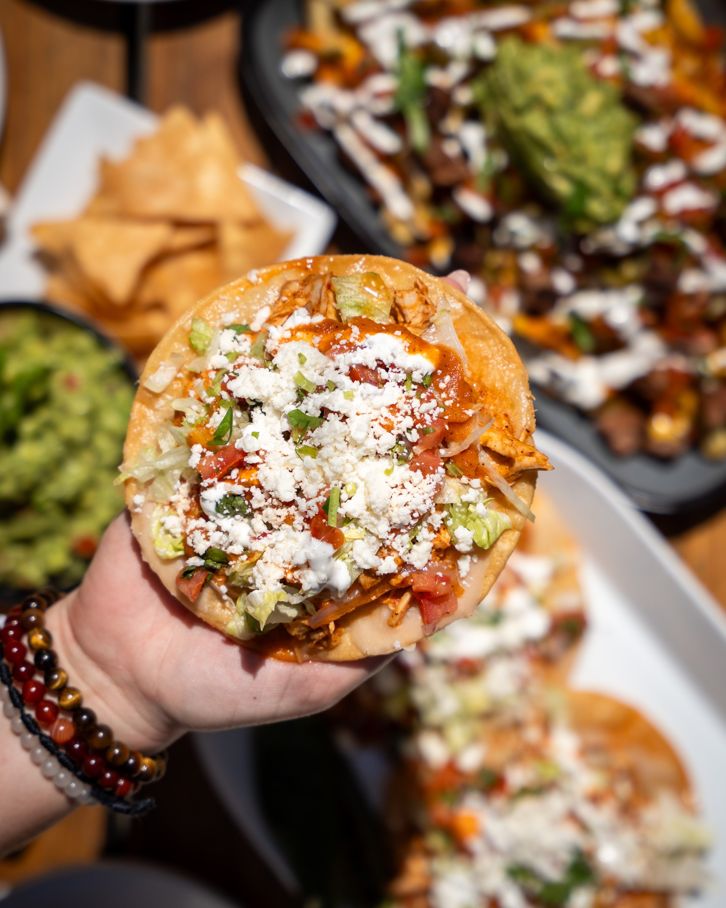 Hand holding a taco filled with lettuce, tomatoes, cheese, and hot sauce, with various dishes in the background