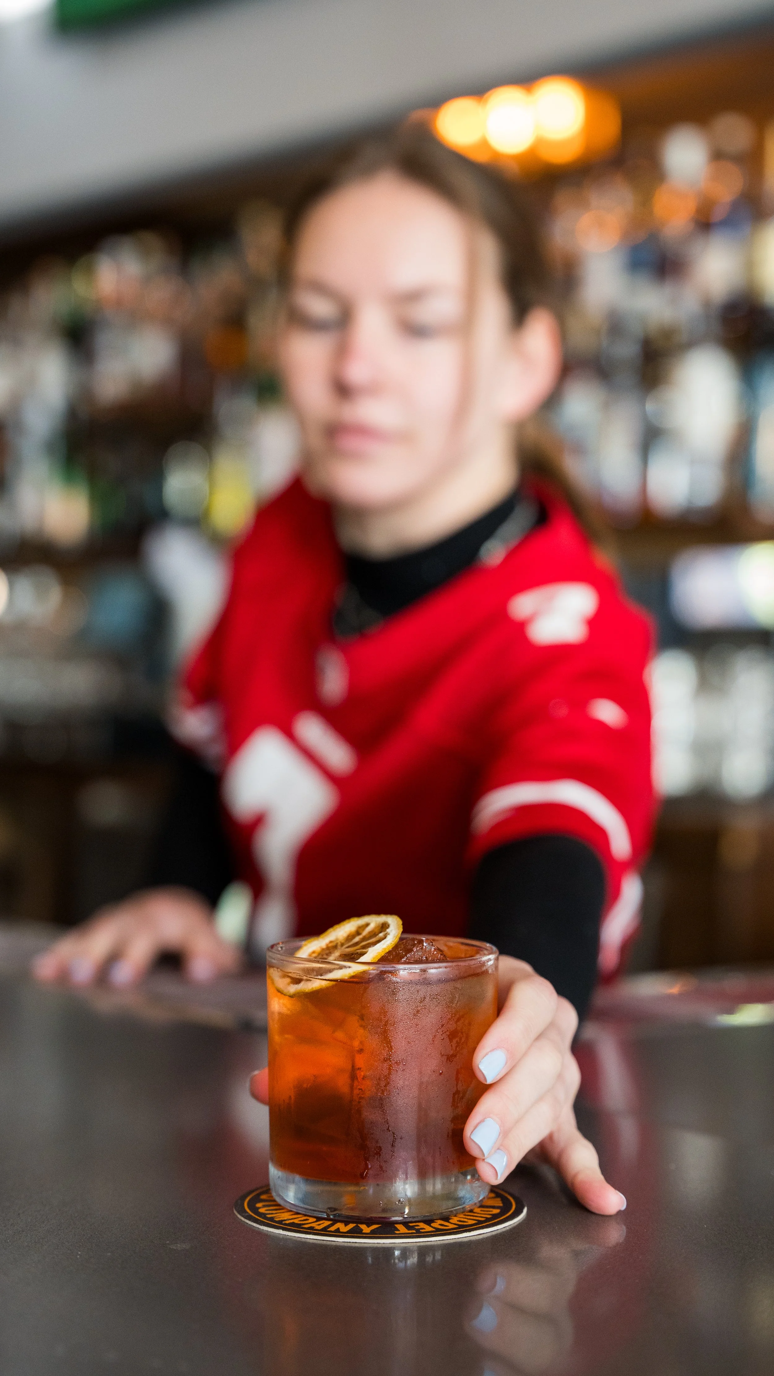 A woman in a red sports jersey serving a drink with lemon slices on the bar counter in a bar or restaurant setting.