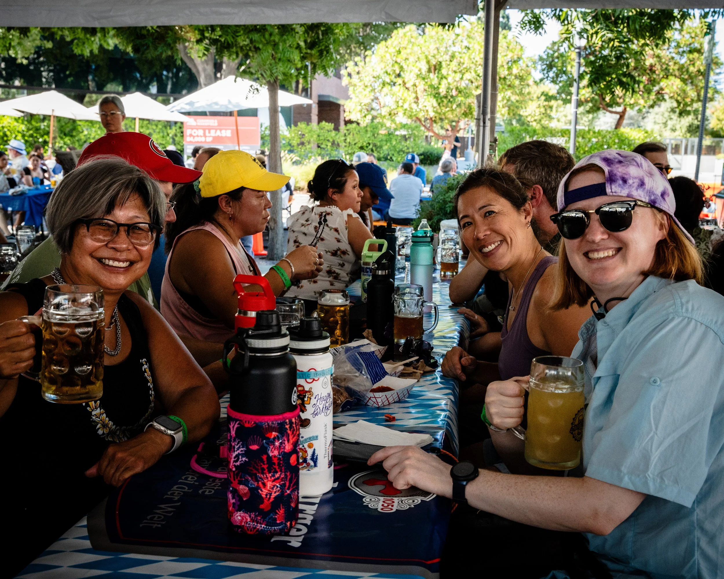Group of people smiling and enjoying drinks at an outdoor event under a tent with trees and other attendees in the background.