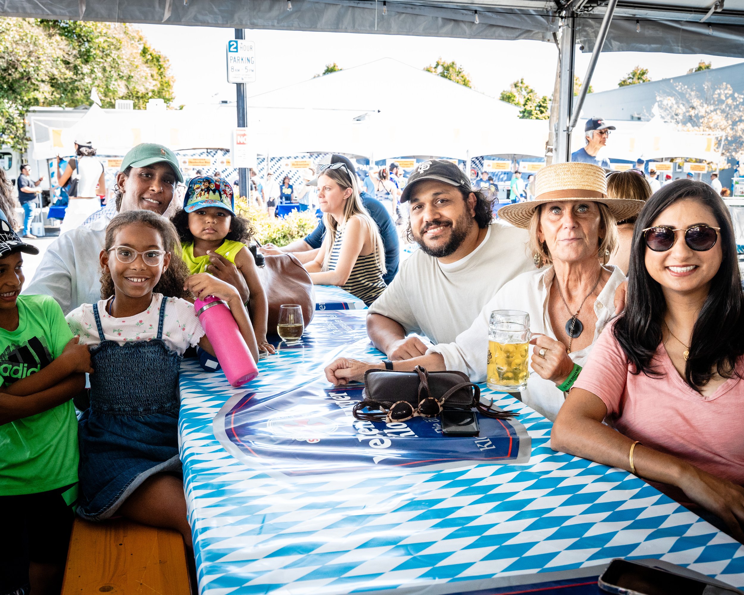 Group of people, including children and adults, sitting at a table under a tent outdoors at a fair or festival. They are smiling, with some holding drinks, and there are vendor booths visible in the background.