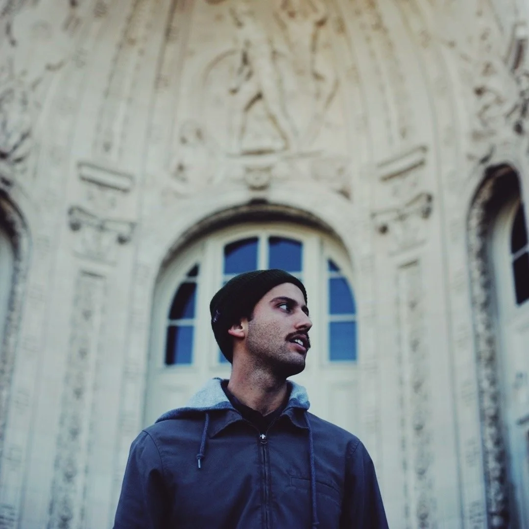 A young man with dark facial hair wearing a black beanie, a dark jacket, and a gray hoodie stands in front of a historic building with detailed architectural carvings and arched windows.