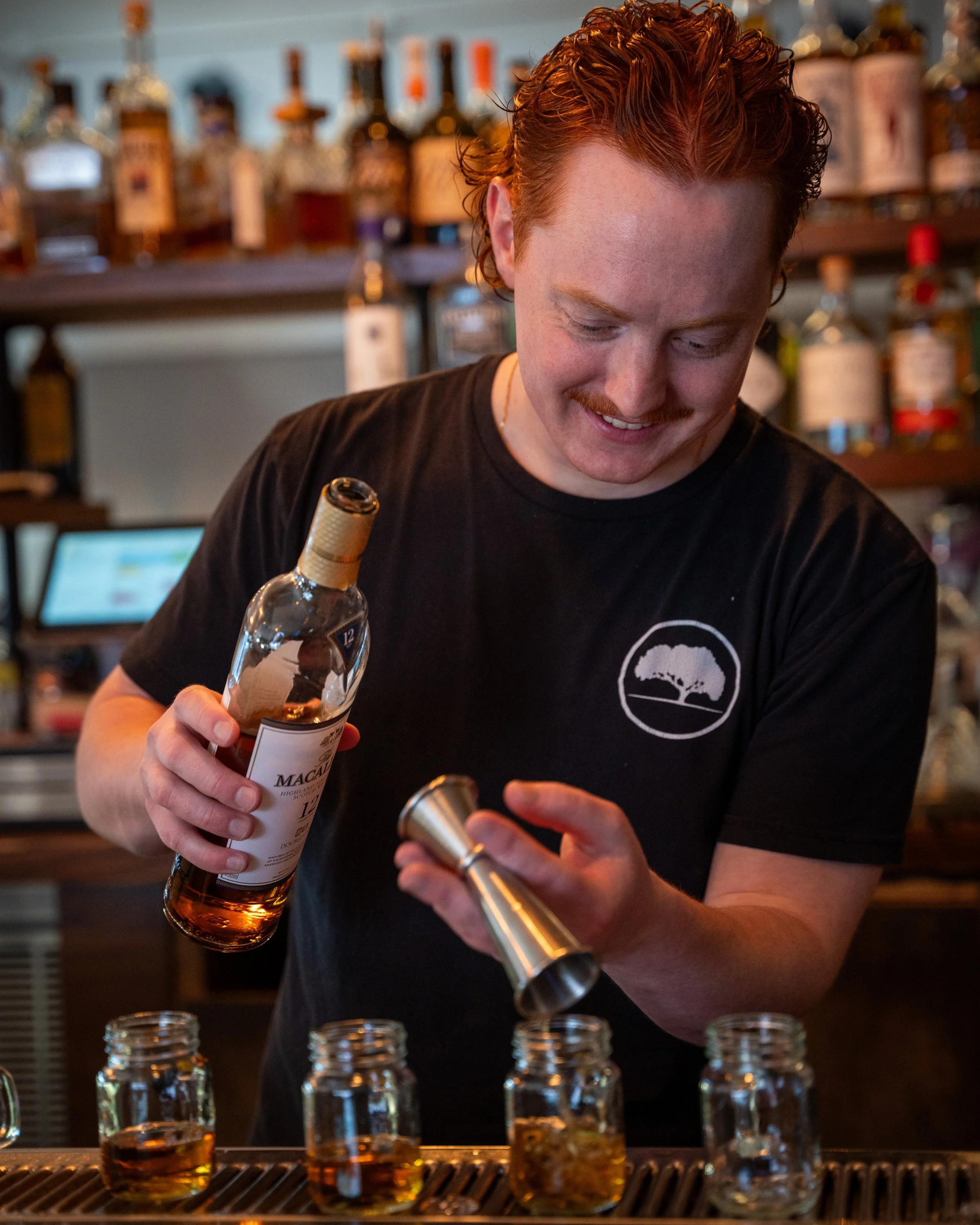 A bartender with red hair and a black t-shirt preparing whiskey drinks at a bar, pouring whiskey from a bottle into small glasses.