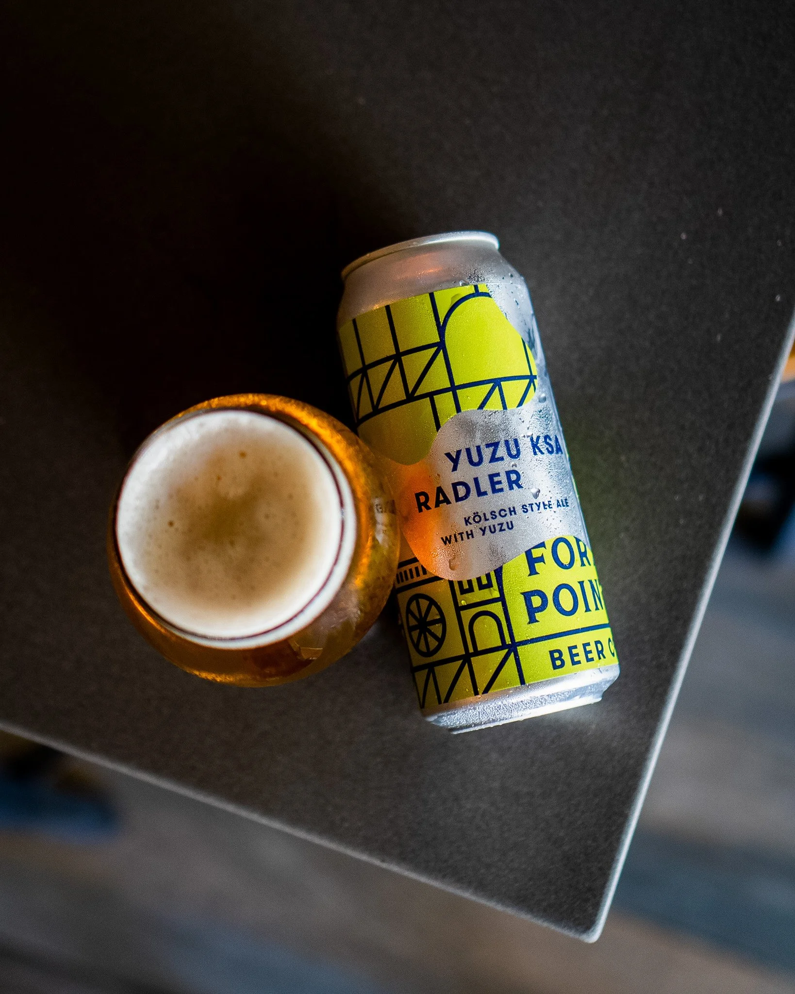 A can of Yuzu KSA Radler beer next to a glass filled with beer, both on a dark surface.