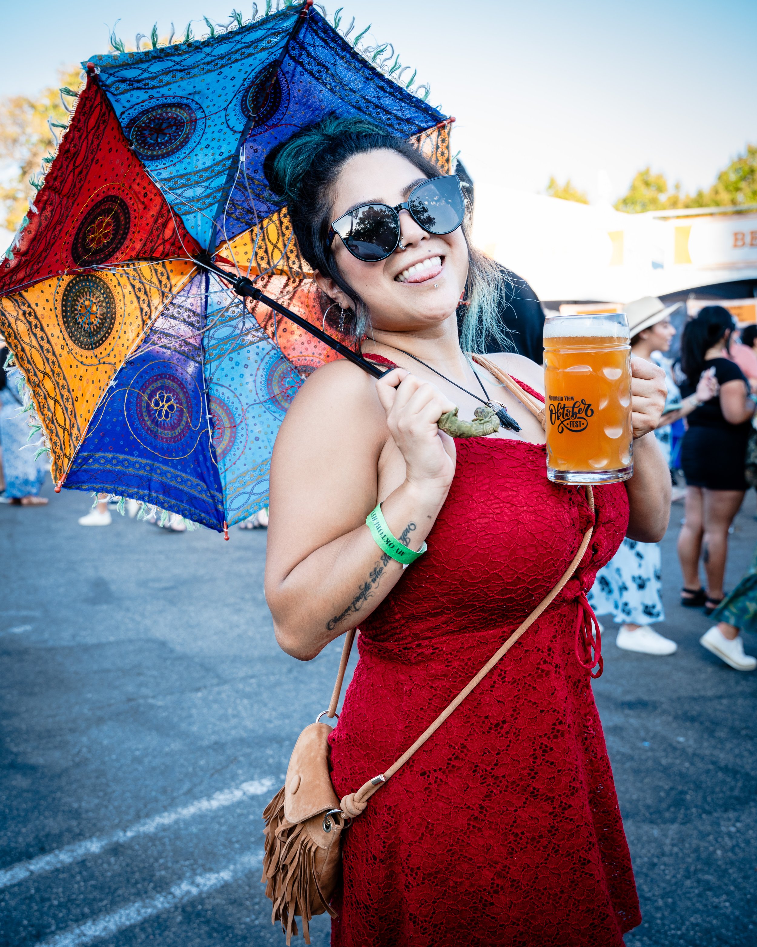 A woman at a festival holding a colorful umbrella and a mug of beer, wearing sunglasses and a red dress, smiling at the camera.