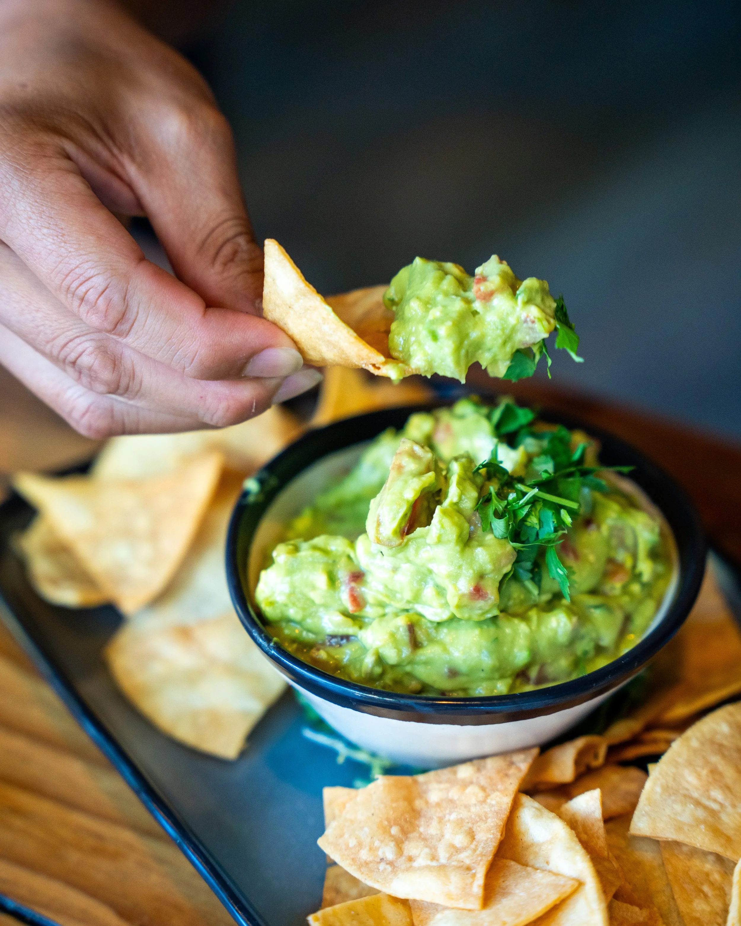 A hand dipping a tortilla chip into a bowl of guacamole with diced tomatoes and herbs, with more chips around the bowl.