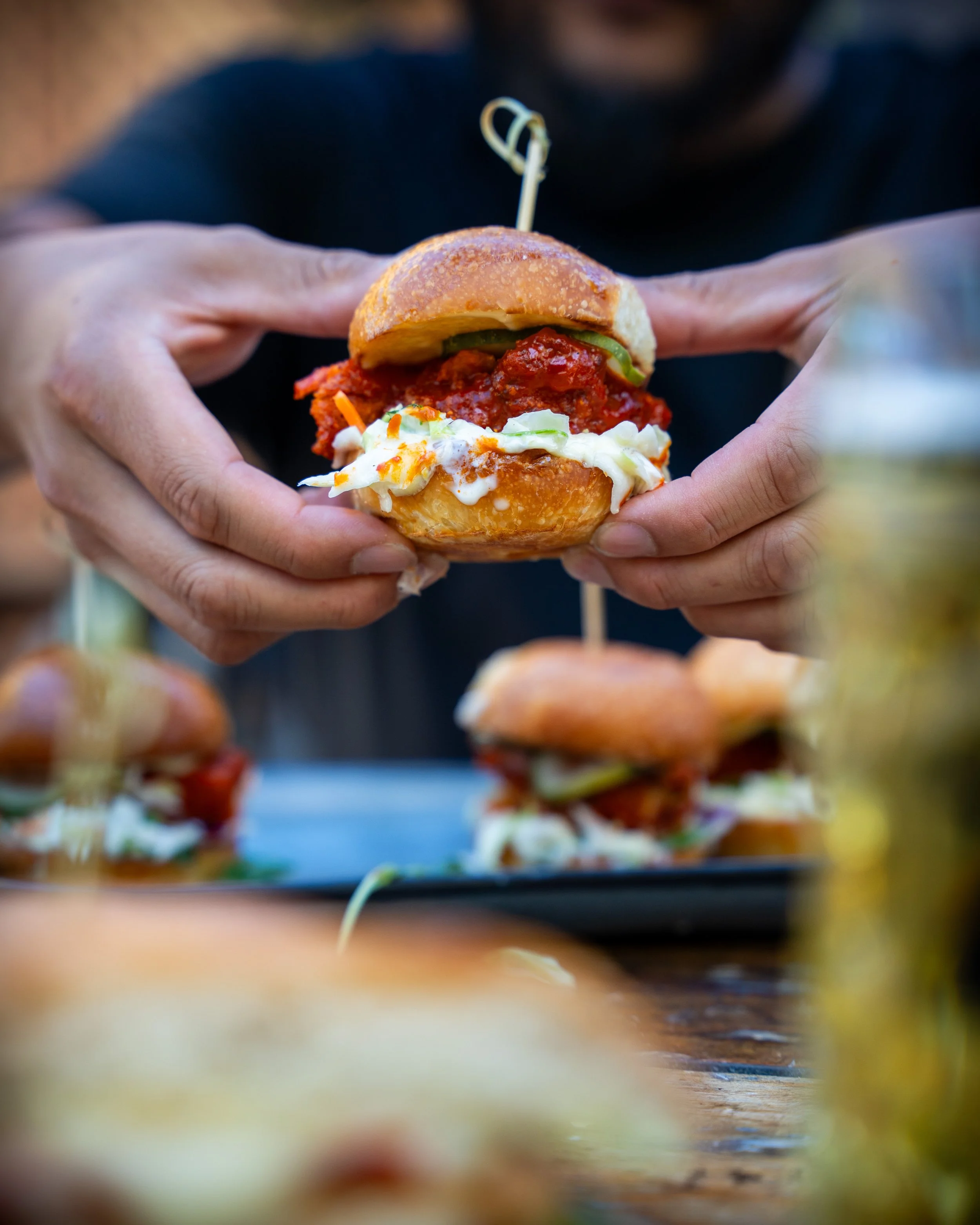 Person holding a small sandwich with fried chicken, pickles, and sauce, with more sandwiches and a drink on the table in the background.