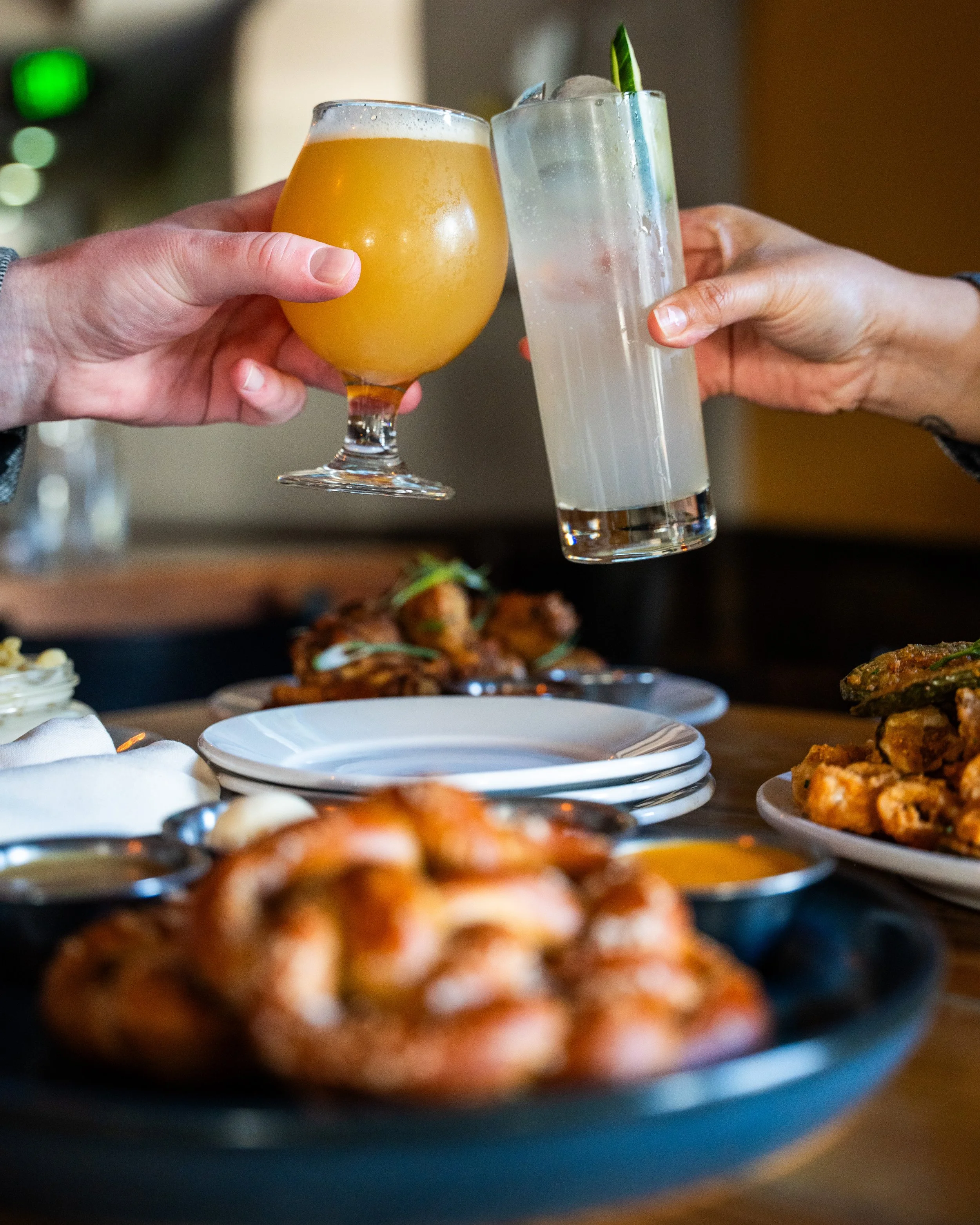 Two people clinking glasses, one with a yellow beer and the other with a clear cocktail with a lime garnish, over a table of various dishes including shrimp, fried chicken, and other appetizers.