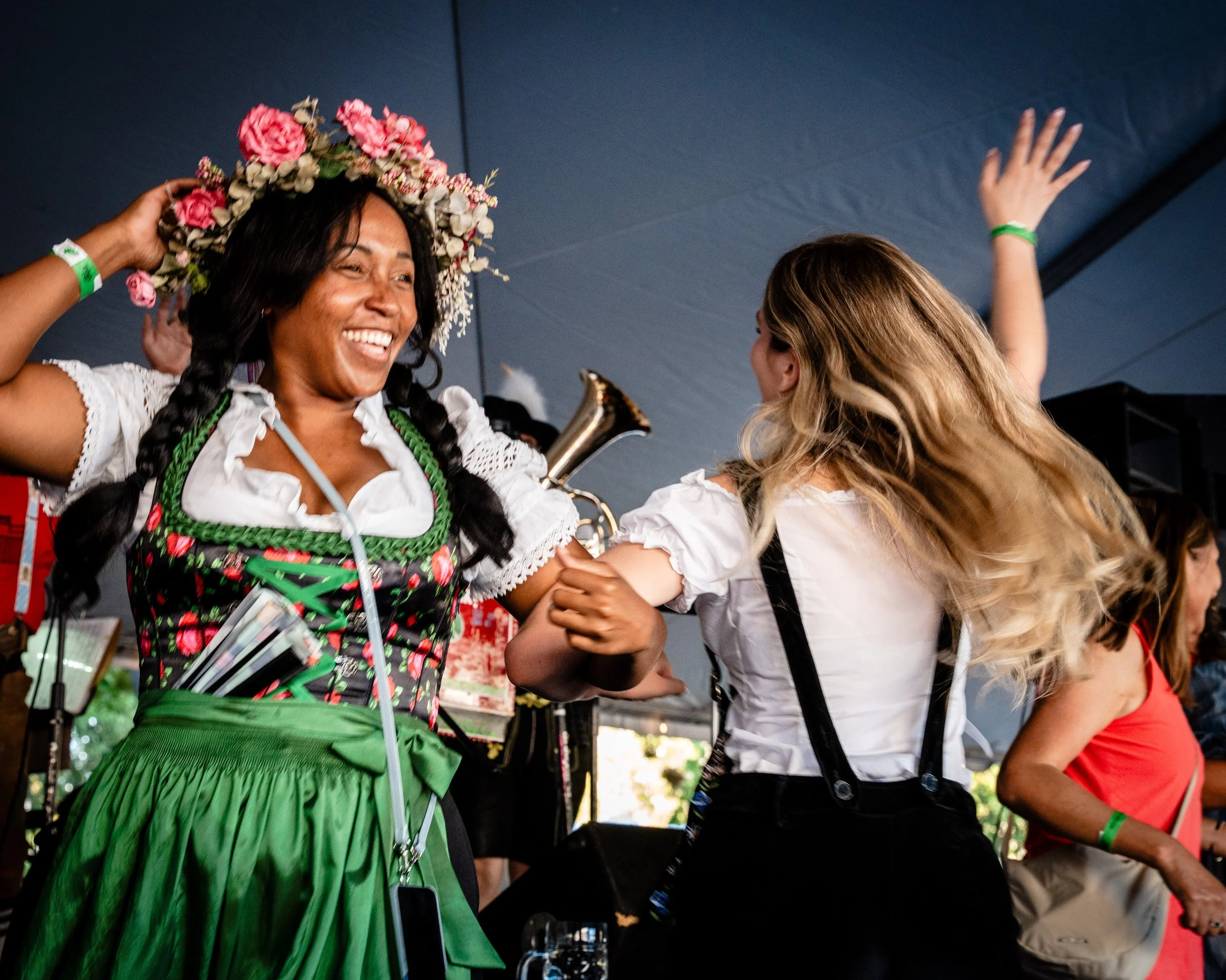 Two women smiling and dancing together, one wearing traditional German attire with a flower crown, and the other in casual clothing, inside a tent at an event.