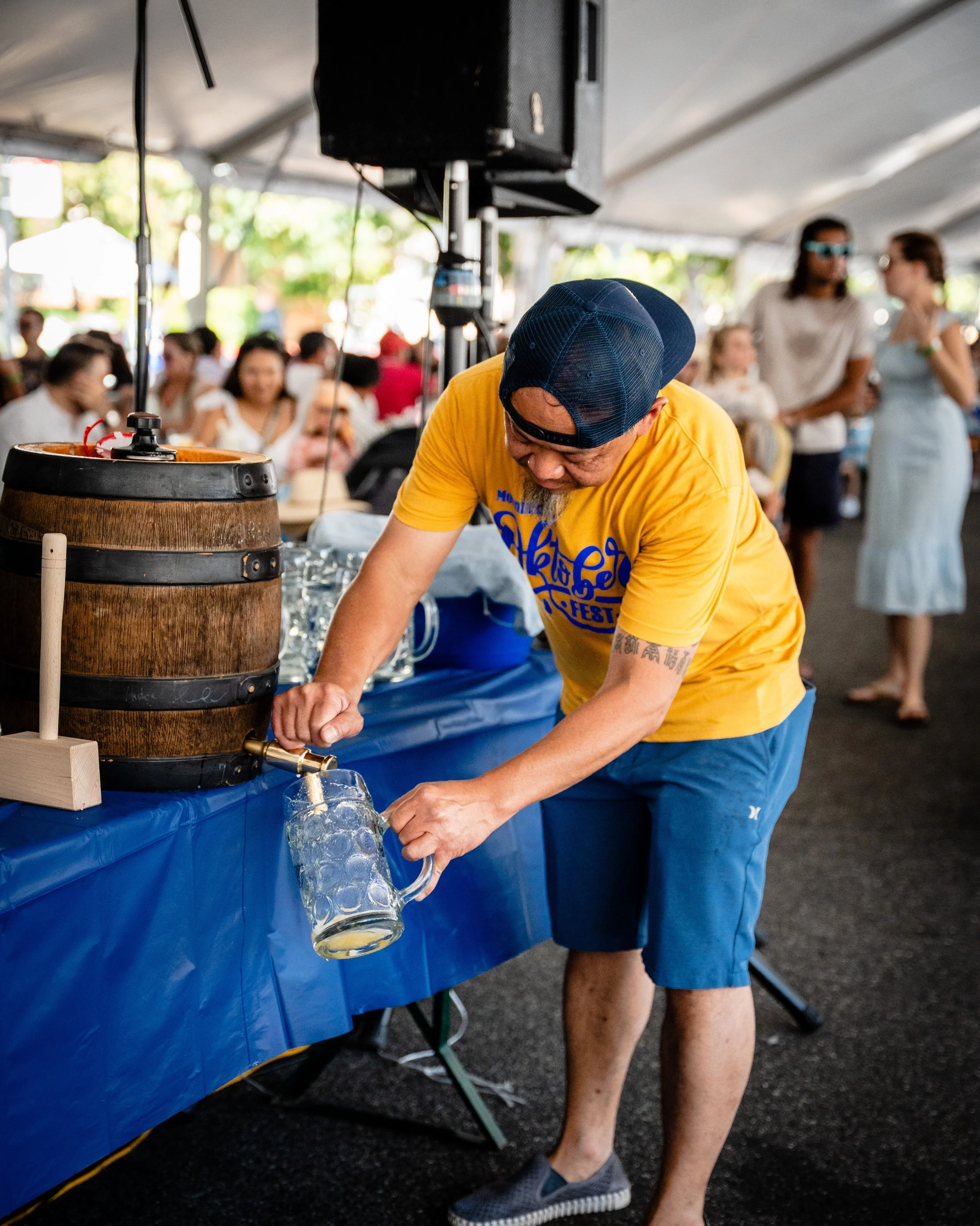 A man pouring beer from a wooden barrel tap into a glass mug at a festival under a large tent, with people socializing in the background.
