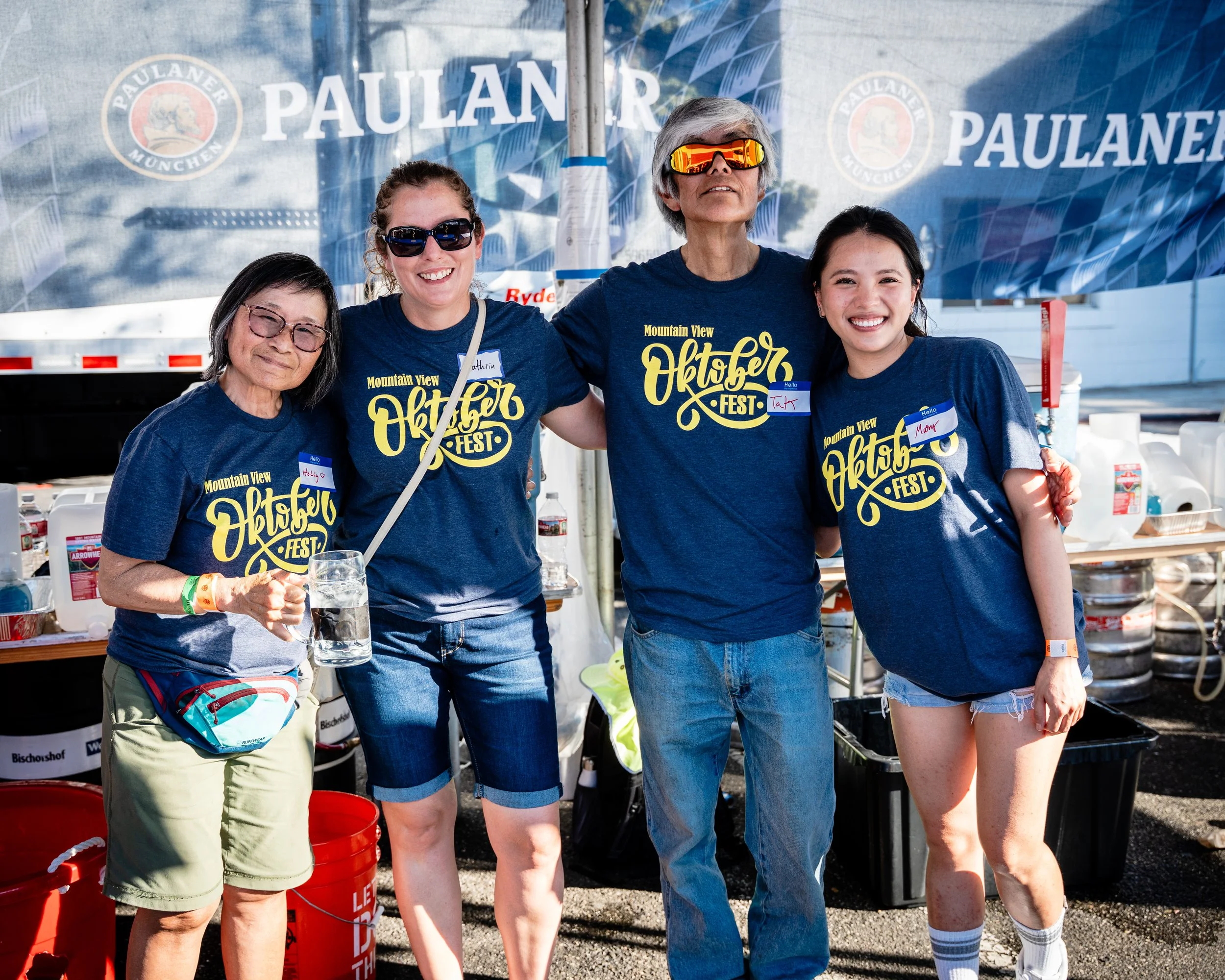 Four people standing together at an outdoor event, wearing matching blue T-shirts with 'Oktober Fest' written on them. They are smiling and posing with arms around each other, with food and drink items visible behind them.