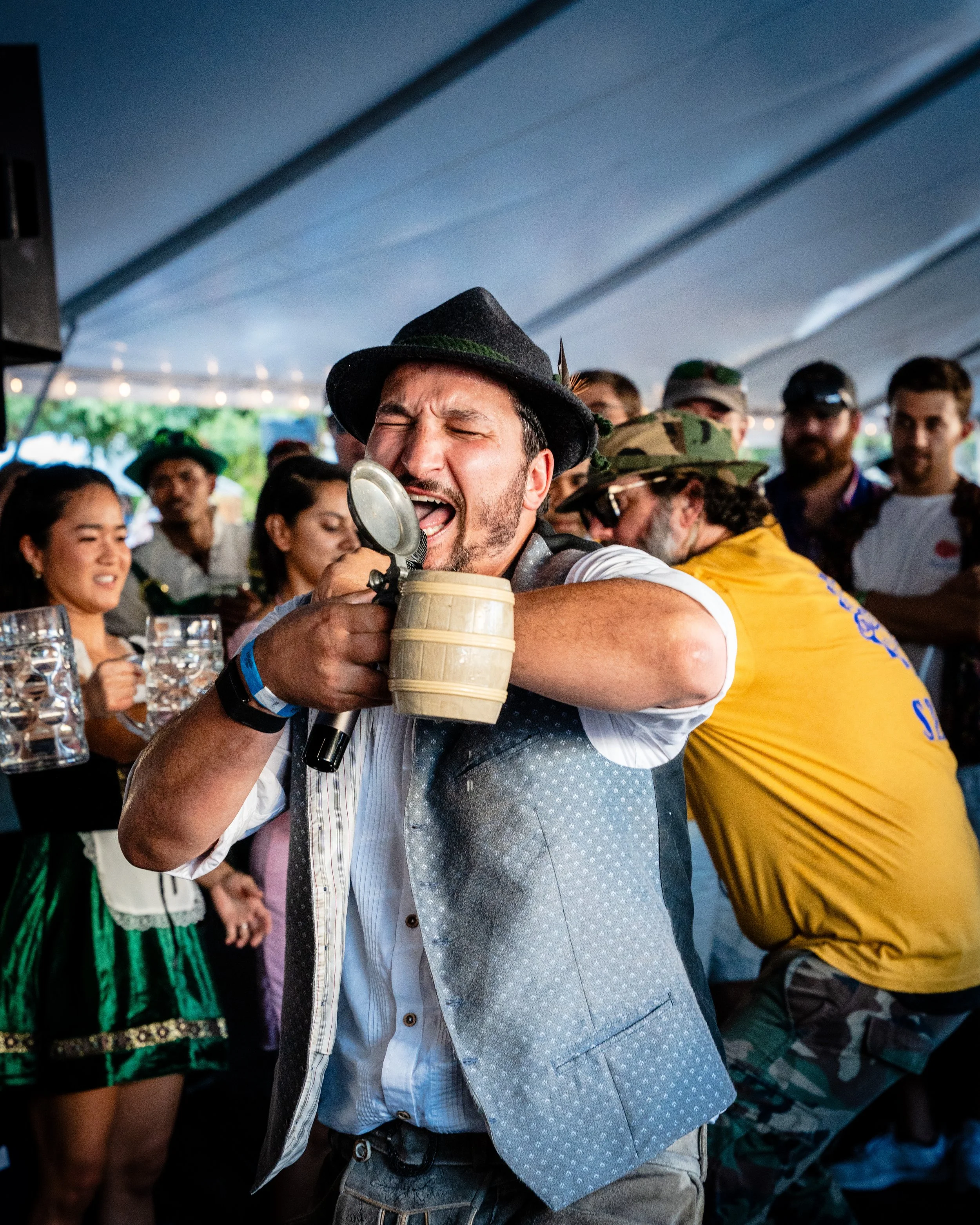 Man singing into a microphone at a lively gathering, wearing a traditional Bavarian hat, with people holding beer mugs and dressed in festive clothing under a tent.