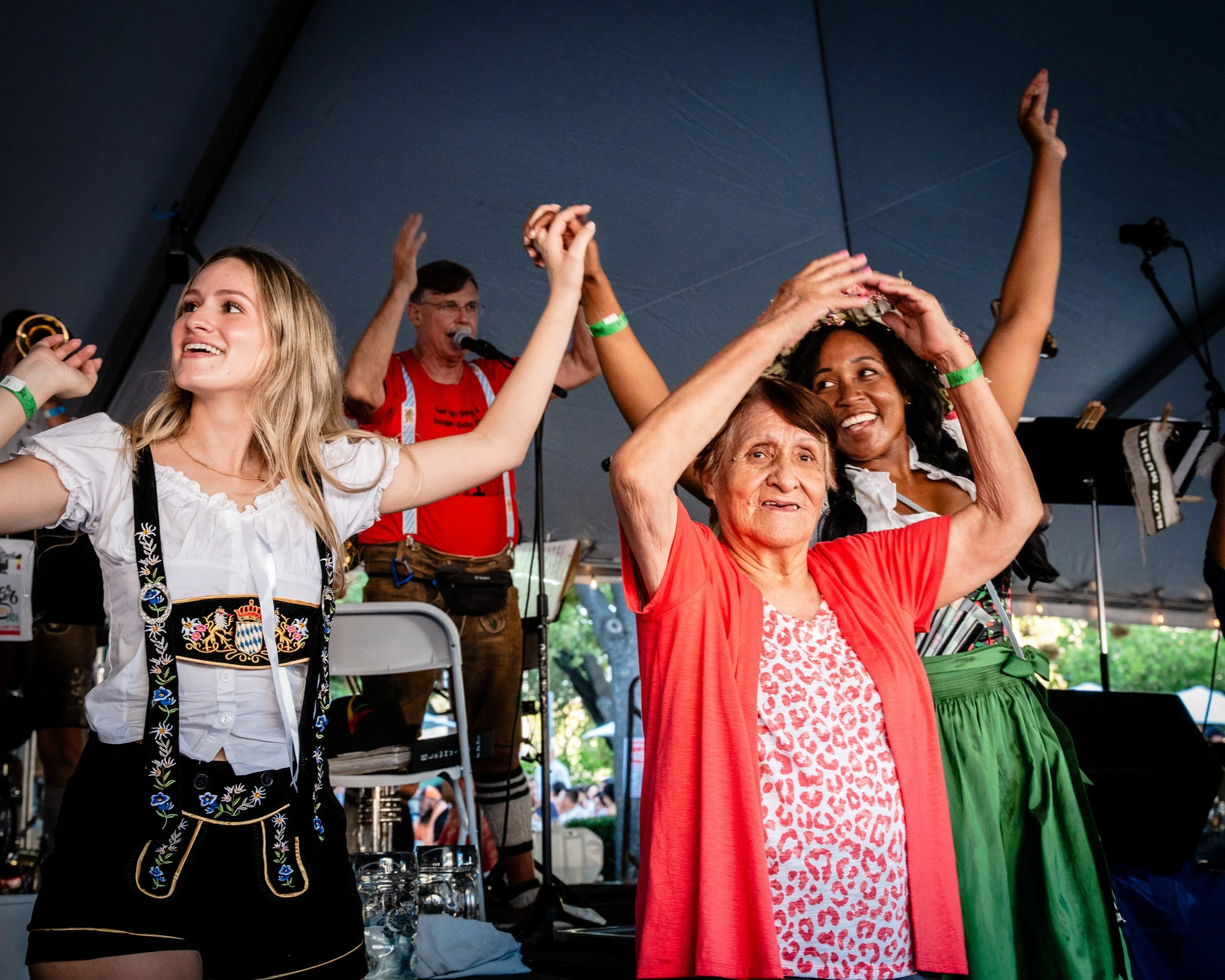 People dancing and celebrating at an event under a large tent, with a woman in front wearing a red cardigan and a woman behind her wearing traditional Bavarian dress, all smiling and enjoying the music.