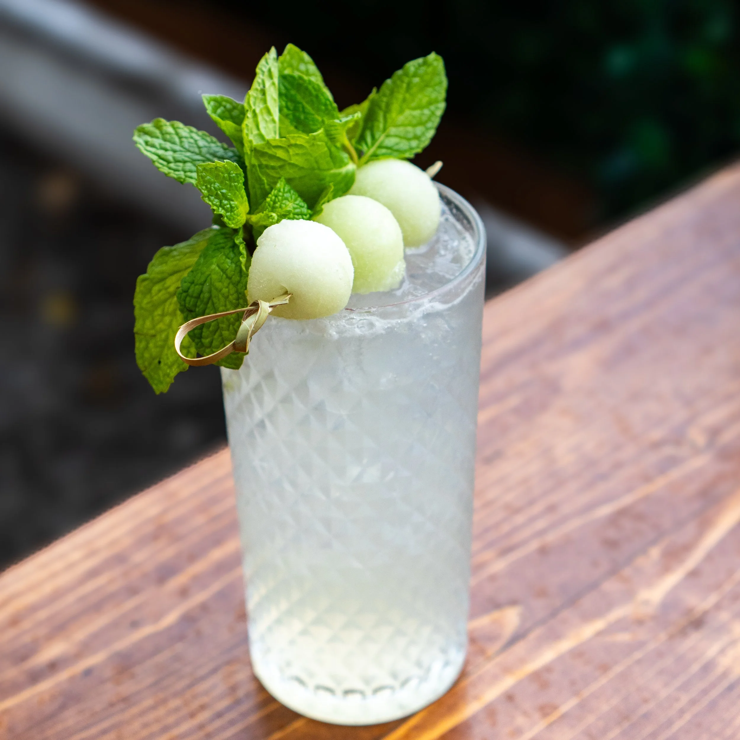 A tall glass of a clear, fizzy drink garnished with mint leaves and green melon balls.
