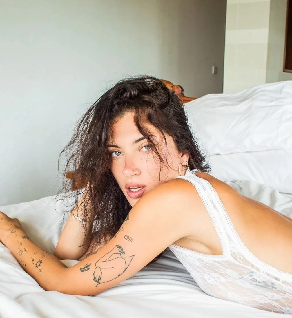 A woman with curly dark hair lying on a bed with white sheets, wearing a white lace tank top, and looking at the camera with an intense expression.