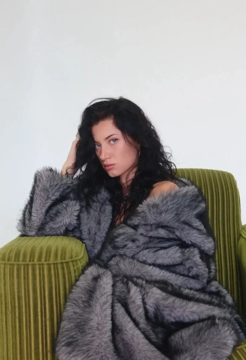 A young woman with long curly black hair and light skin sitting on a green striped armchair, wearing a large gray fur coat, looking at the camera with a serious expression against a plain white wall.