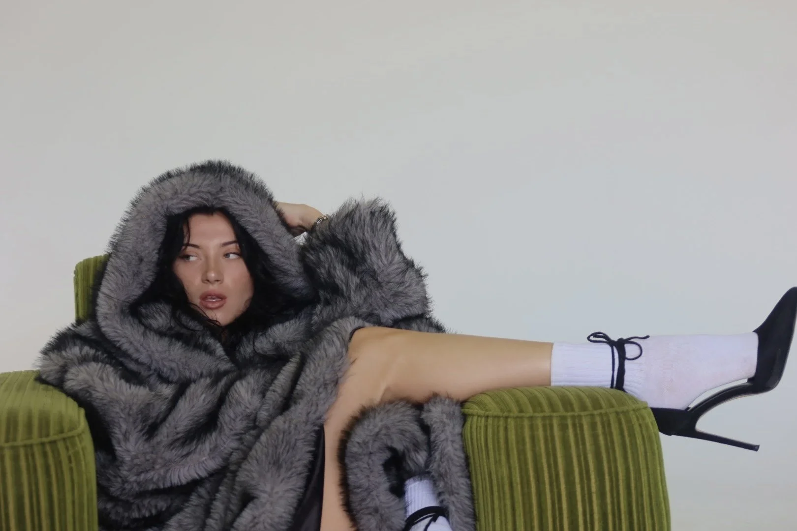 A woman lying on a green couch, wearing a large gray fur coat with a hood, white socks, and black shoes. She has dark hair and is looking to the side with her head resting on her hand.